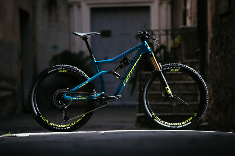 A blue Cannondale mountain bike displayed in a shadowy alley, featuring high-traction tires and a sleek frame design, with sunlight illuminating its contours.