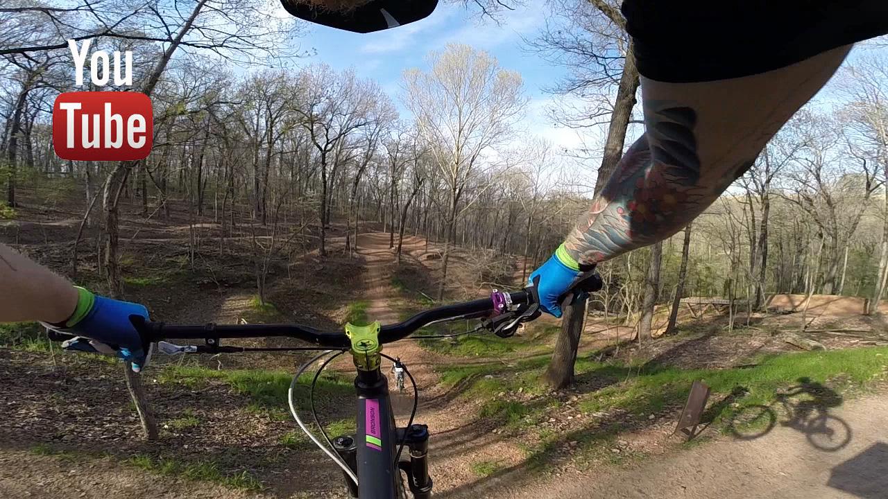 Santa Cruz Bronson: A first-person view of a person mountain biking through a wooded trail, with hands gripping the bike handlebars, showcasing vibrant blue gloves and tattoos on the forearm. The setting features a clear sky and a dirt path winding through the trees, with a silhouette of a cyclist visible in the foreground. The image includes a YouTube logo in the top left corner.