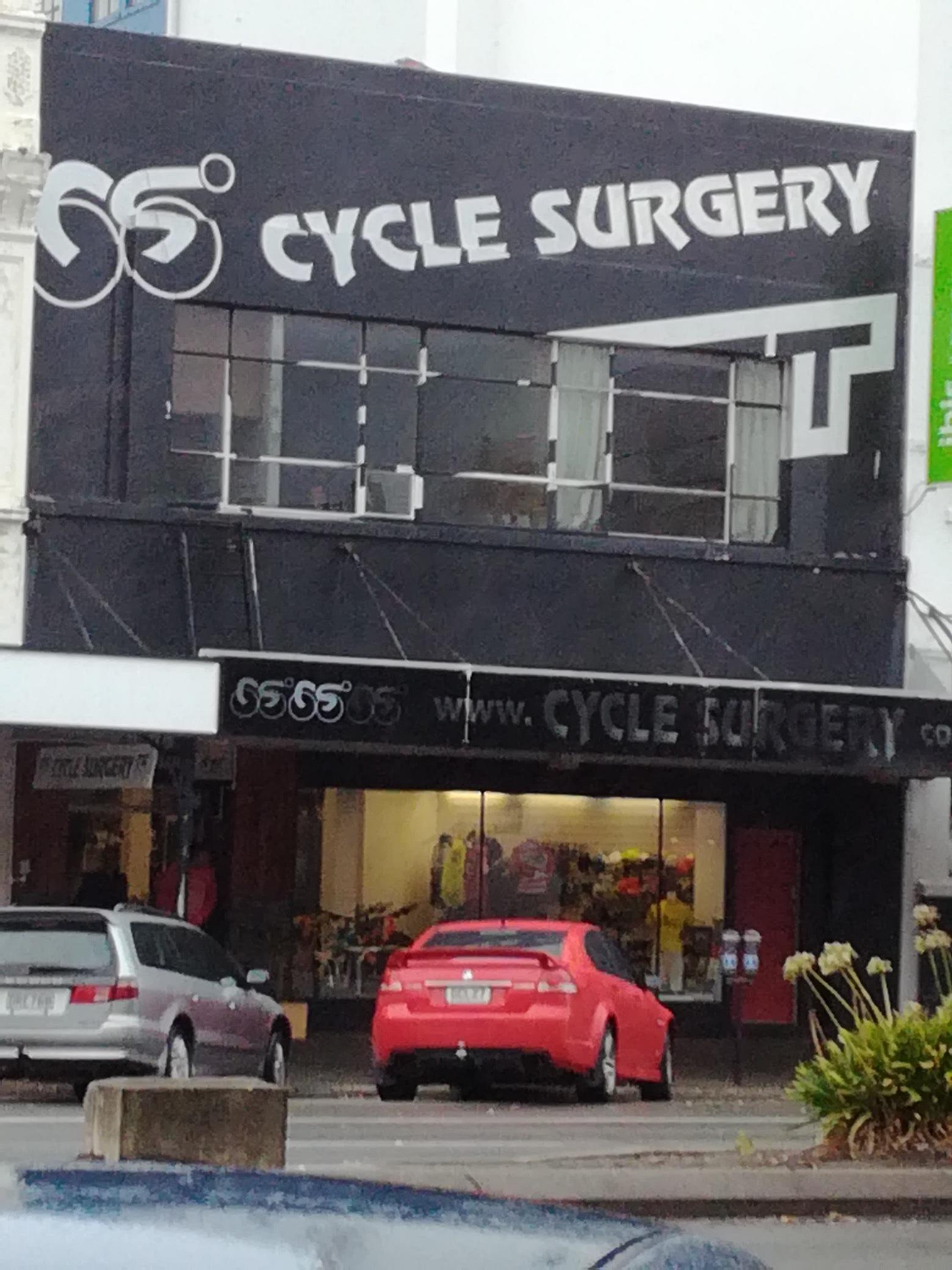 Exterior view of a store named "Cycle Surgery," featuring a large black sign with white lettering. The shop displays various cycling gear and accessories in the windows. A silver car is parked next to a bright red car in the foreground, along with some greenery visible at the bottom of the image. The overall atmosphere suggests a bustling urban setting.