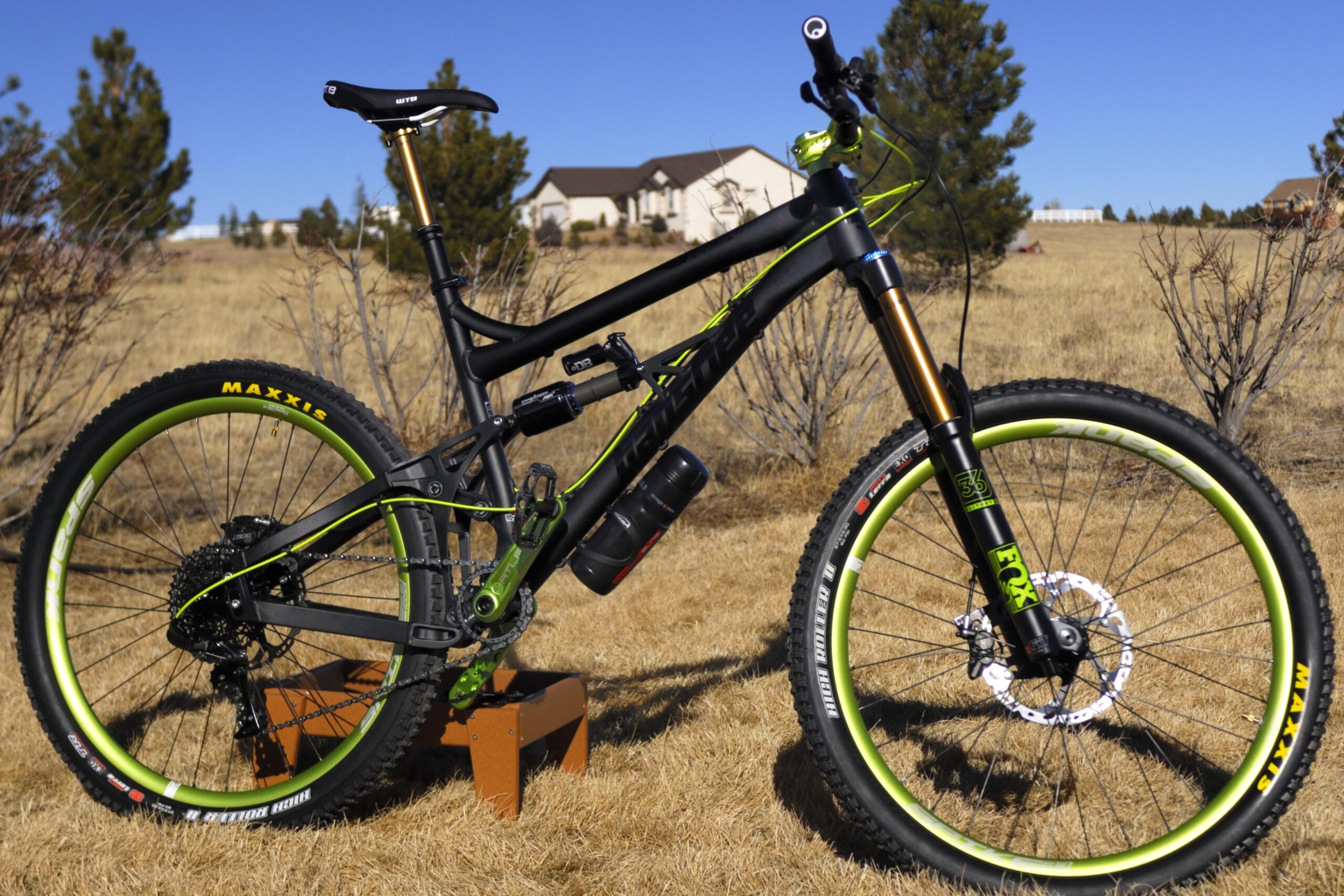 Banshee Rune: A black and green mountain bike rests on a wooden stand in a grassy field. The bike features wide Maxxis tires, a dropper seat post, and a water bottle mounted on the frame. In the background, there are sparse bushes and a few distant houses under a clear blue sky.
