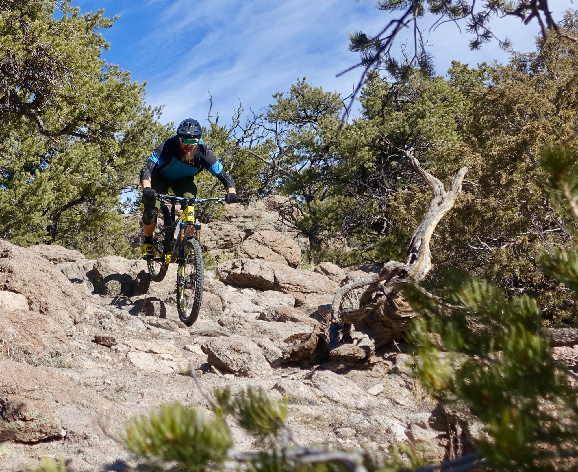 A mountain biker wearing a black helmet and sunglasses descends a rocky trail surrounded by trees. The rider is captured mid-jump over a boulder, showcasing the excitement and skill involved in mountain biking on rugged terrain. The blue sky and greenery add to the outdoor adventure atmosphere. Unkle Nazty mountain bike trail.