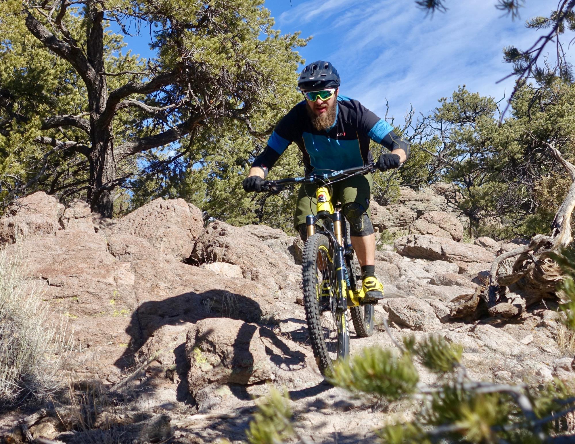 A mountain biker navigating a rocky trail surrounded by trees on a sunny day. The cyclist is wearing a helmet, sunglasses, and protective gear, and is actively riding over the uneven terrain. Unkle Nazty mountain bike trail.