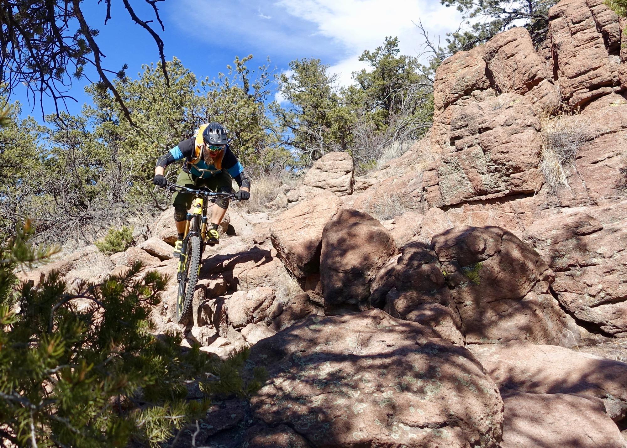 A mountain biker navigating a rocky trail surrounded by trees and blue skies. The rider is leaning forward on the bike, showcasing an action shot as they tackle the uneven terrain. The scene captures the essence of outdoor adventure and sport in a natural setting. Unkle Nazty mountain bike trail.