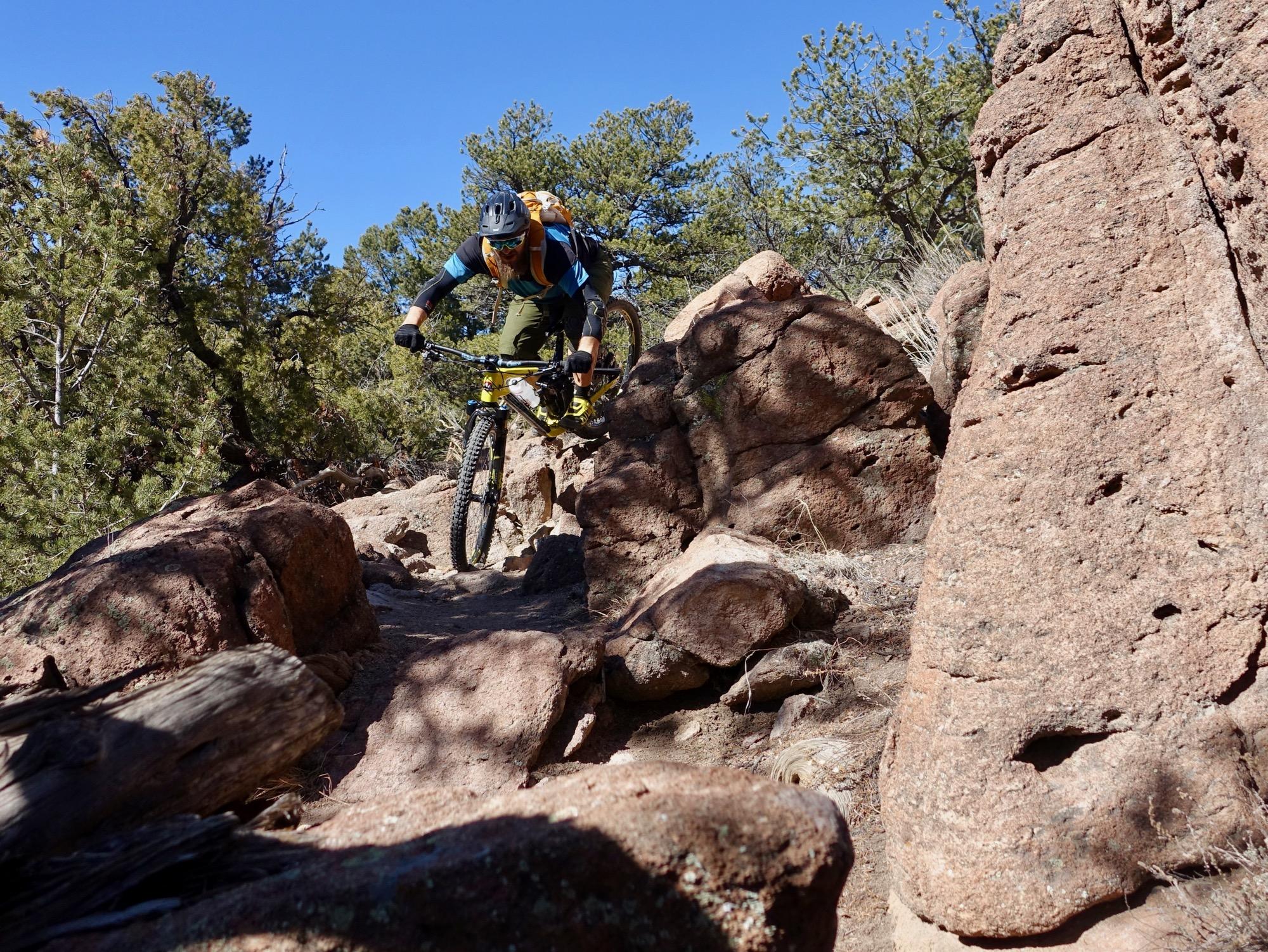 A mountain biker navigating a rocky trail surrounded by trees under a clear blue sky. The rider is leaning forward on their bike, displaying concentration while tackling the uneven terrain. Unkle Nazty mountain bike trail.