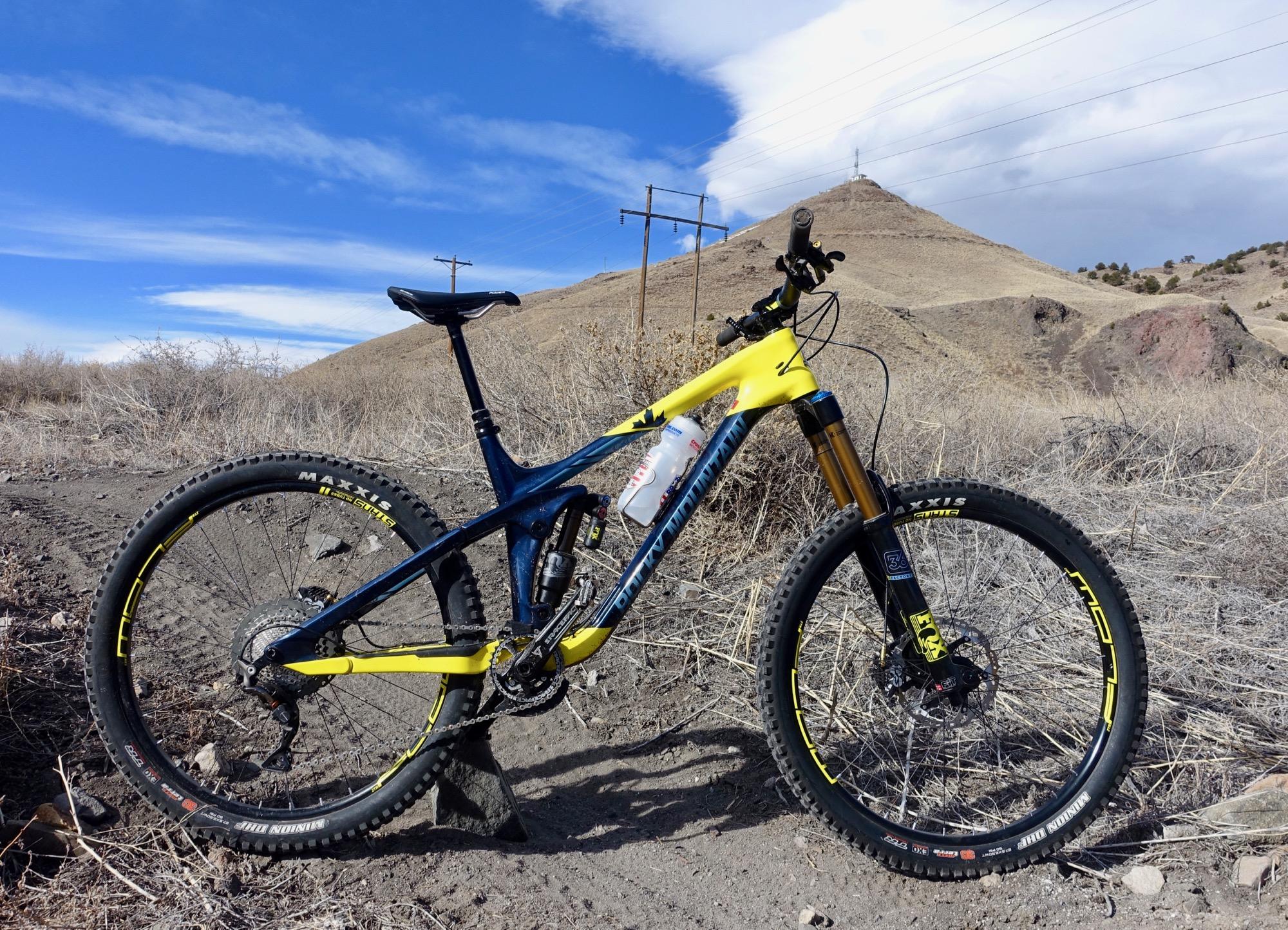 Rocky Mountain Slayer 790 MSL: A brightly colored mountain bike positioned on a dirt trail, with dry, sparse vegetation and a hilly landscape in the background. The sky is clear with a few clouds, and power lines can be seen stretching across the scene.