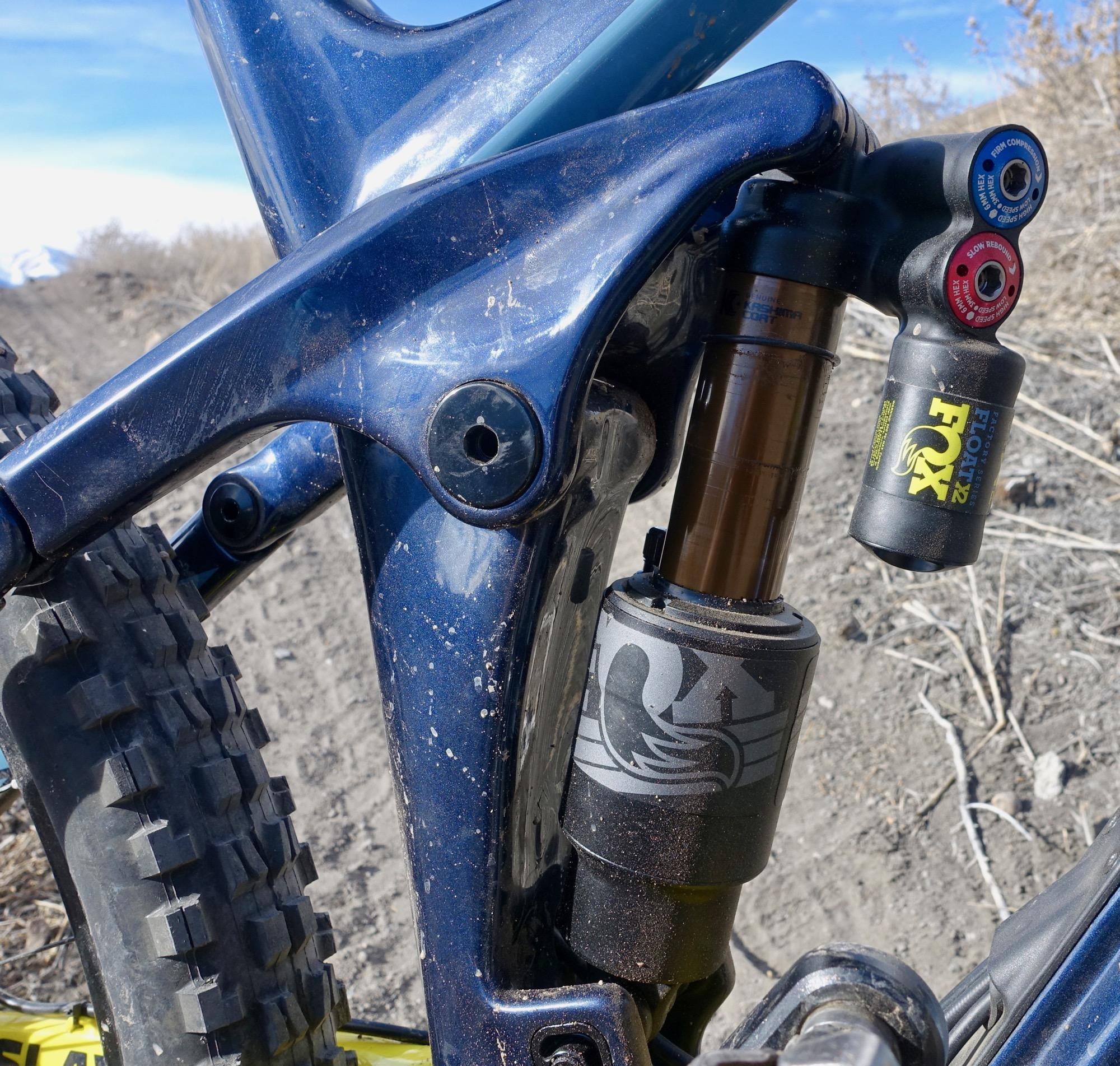 Rocky Mountain Slayer 790 MSL: Close-up image of a mountain bike's rear suspension system, featuring a blue frame, a shock absorber with adjustable dials, and tire treads visible in the background. The setting appears to be an outdoor environment with dirt and sparse vegetation.
