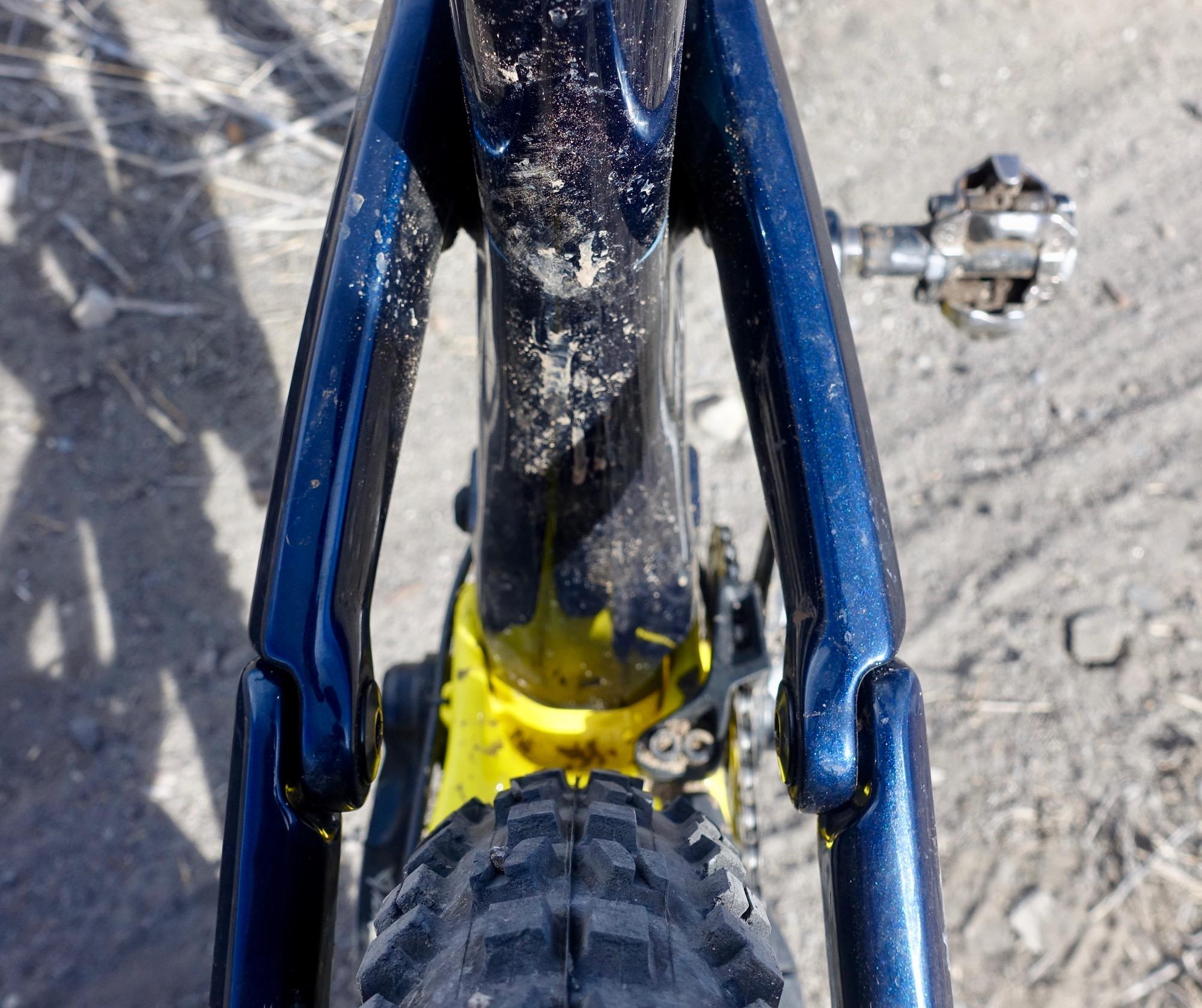 Rocky Mountain Slayer 790 MSL: Close-up view of a mountain bike frame, focusing on the connection between the rear triangle and the main tube. The frame is a glossy blue with visible dirt and dust, highlighting its use in outdoor activities. A portion of the bike's yellow underside is also visible, along with a textured tire tread. The background features a gravel surface, indicating a natural riding environment.