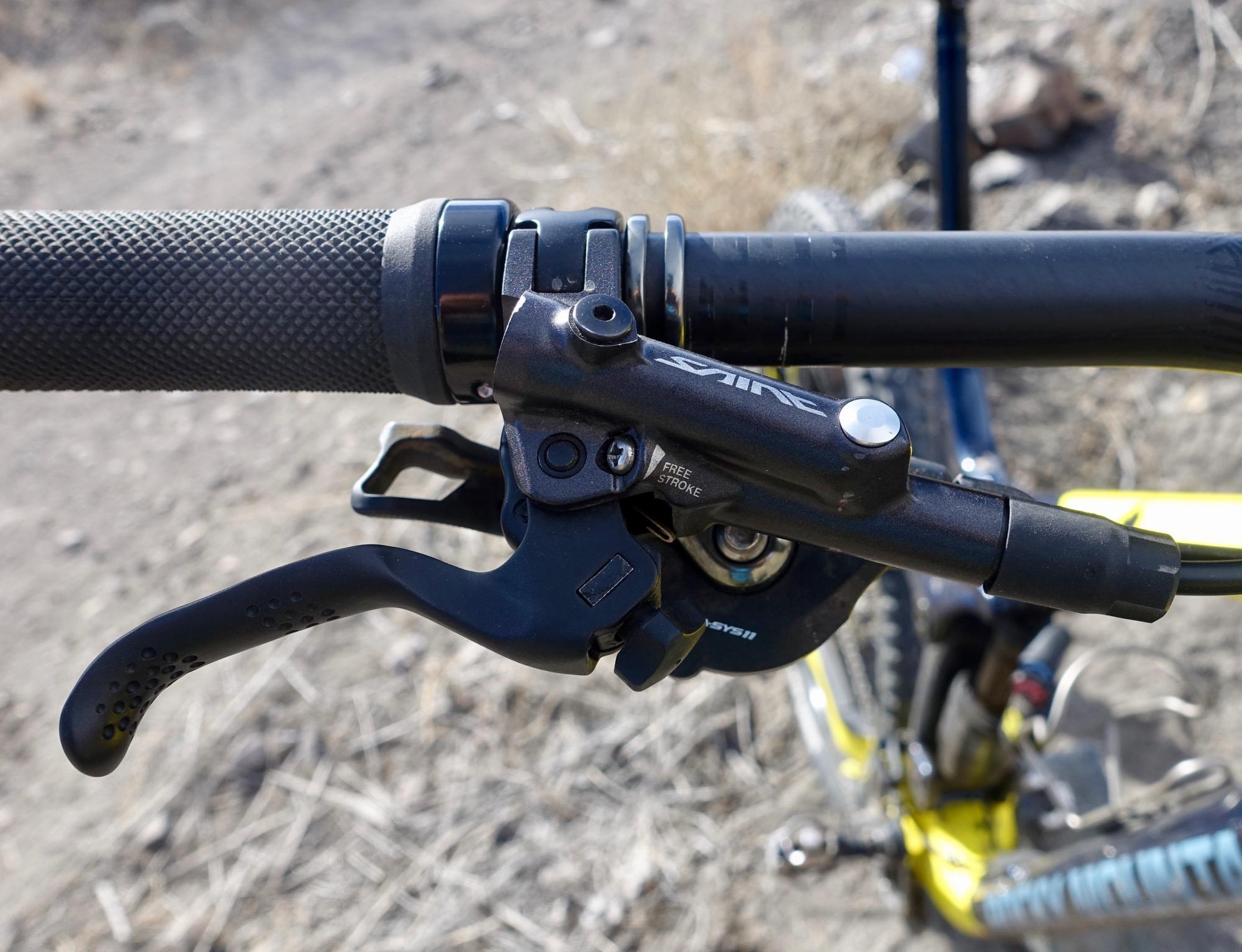 Rocky Mountain Slayer 790 MSL: Close-up view of a bicycle handlebar featuring a textured grip and a hydraulic brake lever, showing the lever's mechanics and branding. The background is a blurred outdoor setting with dry vegetation.