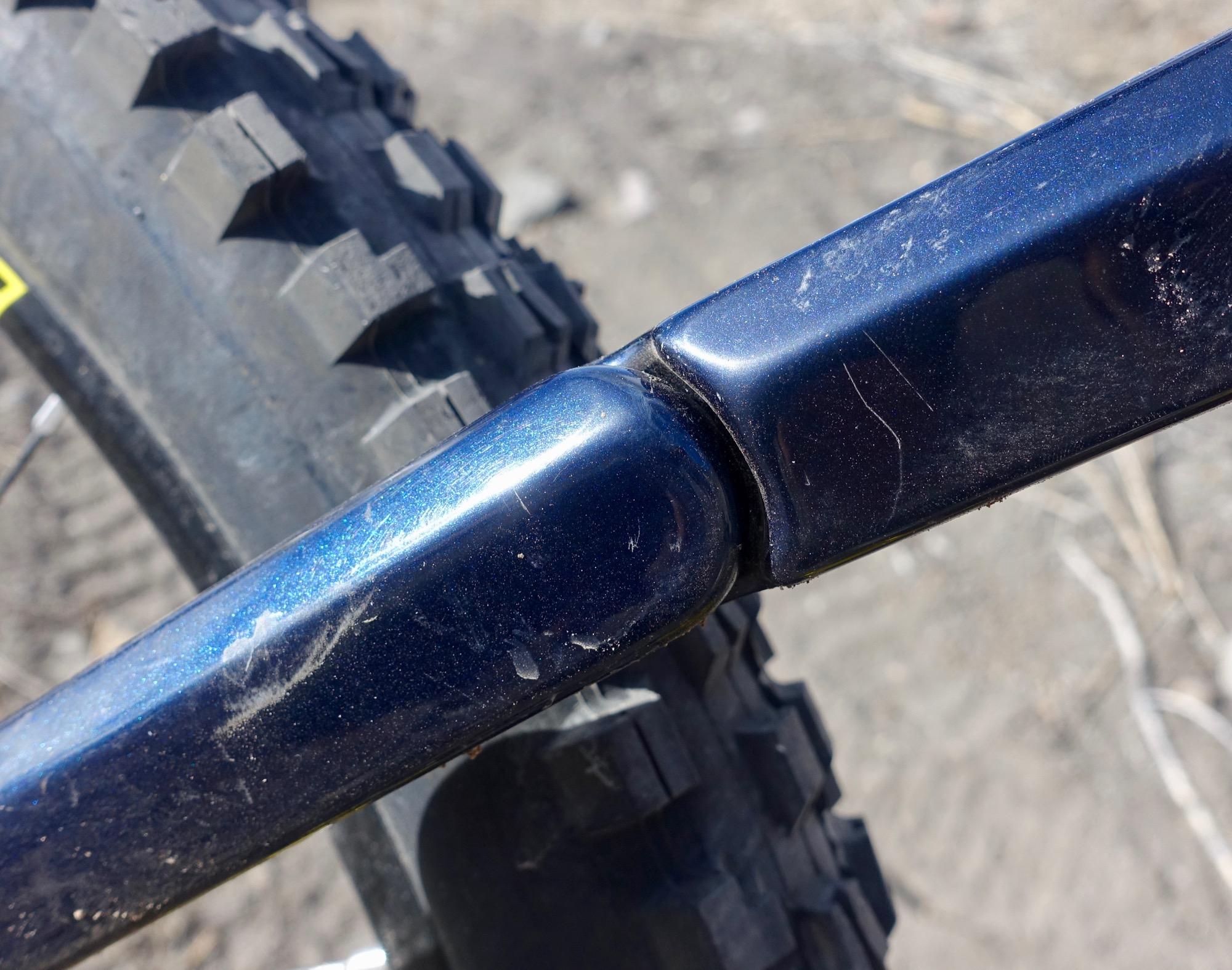 Rocky Mountain Slayer 790 MSL: Close-up image of a blue mountain bike frame showing a joint and part of the tire, with visible dirt and scratches on the frame. The background features a textured, sandy surface.