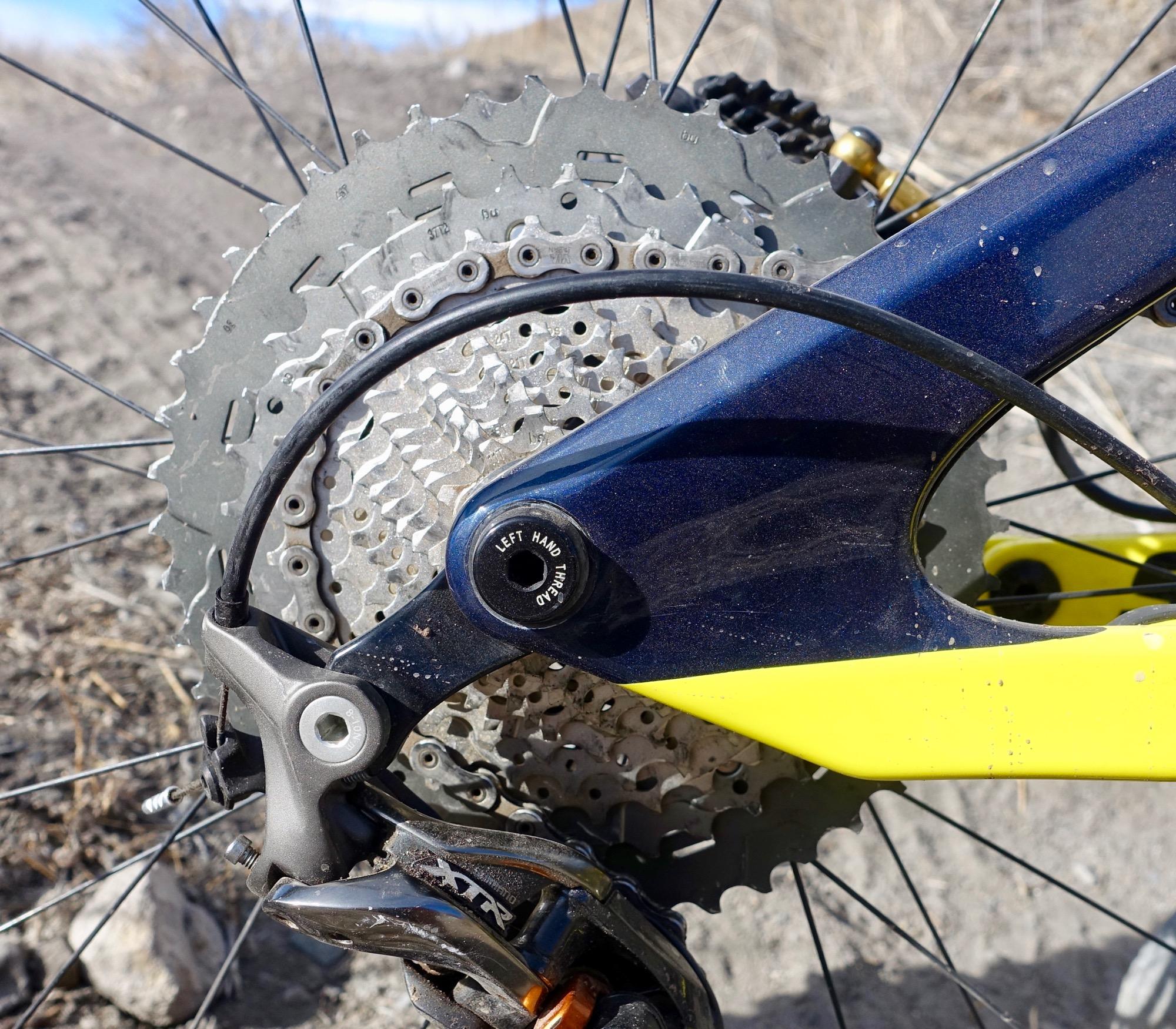Rocky Mountain Slayer 790 MSL: Close-up view of a bicycle's rear derailleur, chain, and cassette, showcasing the metal components. The background features a gravel surface, suggesting an outdoor setting. The bicycle frame is partially visible, displaying a blue and yellow color scheme.