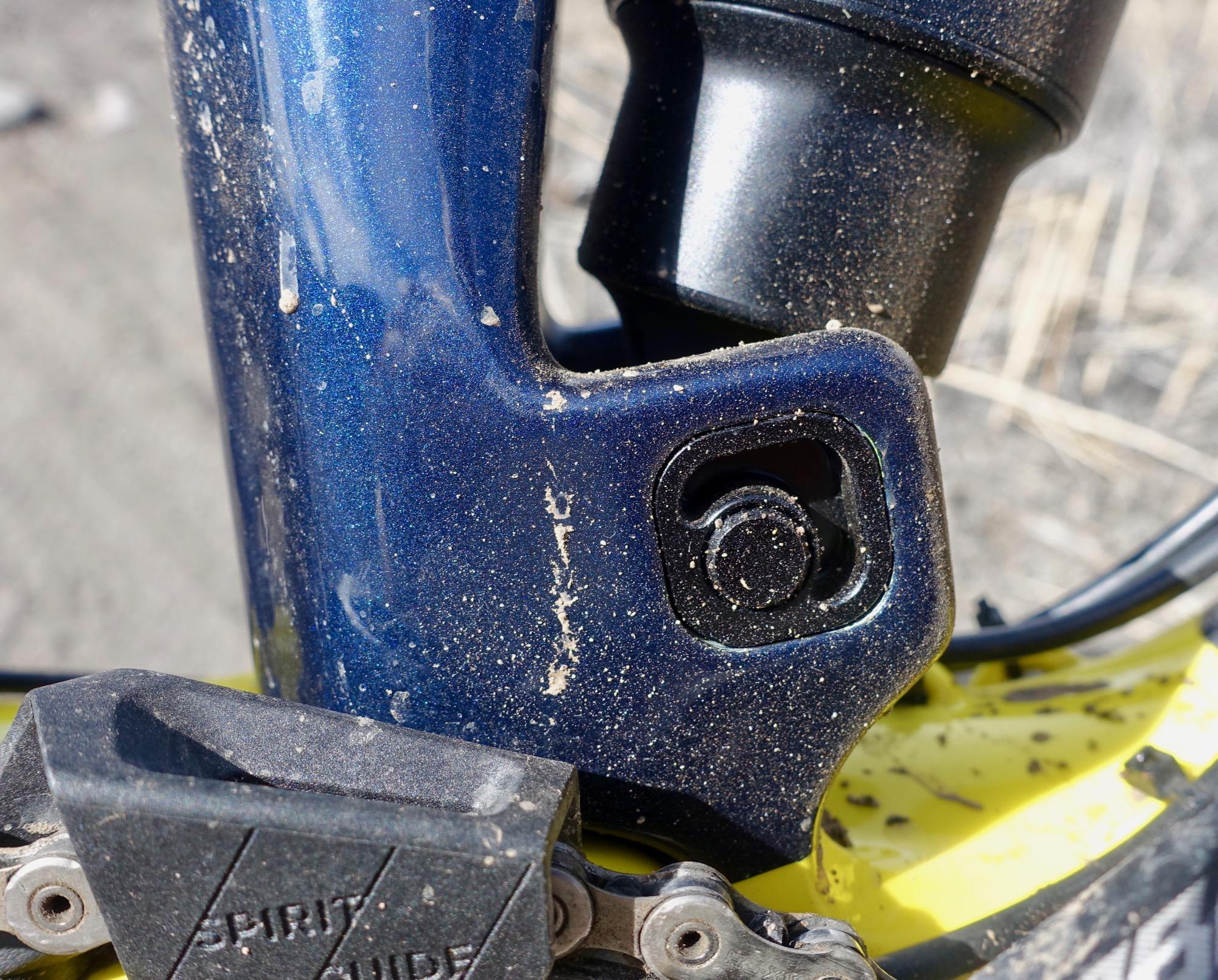 Rocky Mountain Slayer 790 MSL: Close-up image of a bicycle frame with a dark blue finish, showing dirt and dust accumulation. The focus is on the lower part of the frame where the shock mount is located, revealing details of the metallic components and the texture of the paint. A portion of the bike pedal and chain is visible at the bottom of the image.