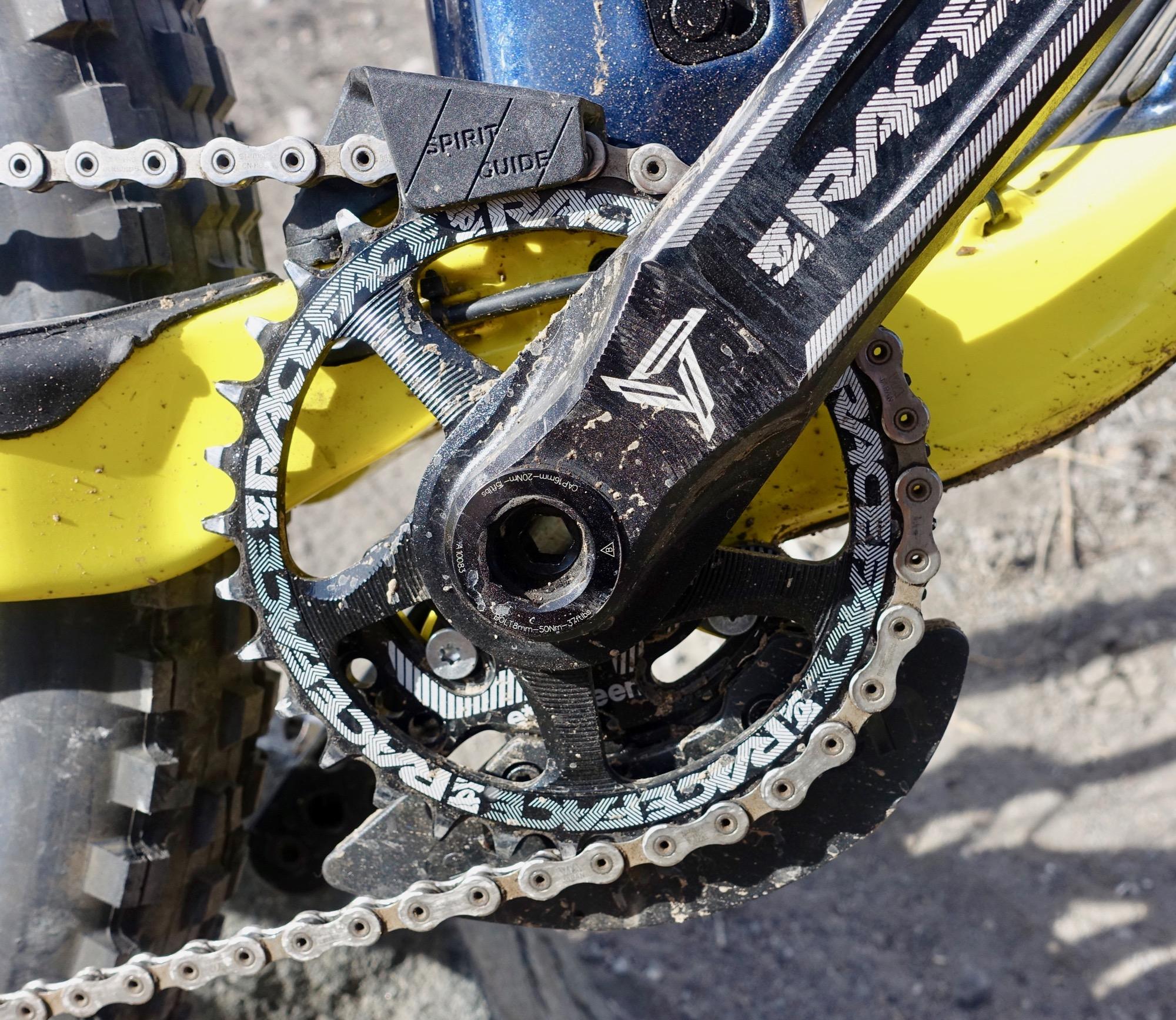 Rocky Mountain Slayer 790 MSL: Close-up view of a mountain bike's drivetrain, showcasing the chainring, chain, and crankset. The components are slightly muddy, indicating off-road usage, with a yellow frame partially visible in the background. The image captures the intricate details of the bike's mechanical parts.