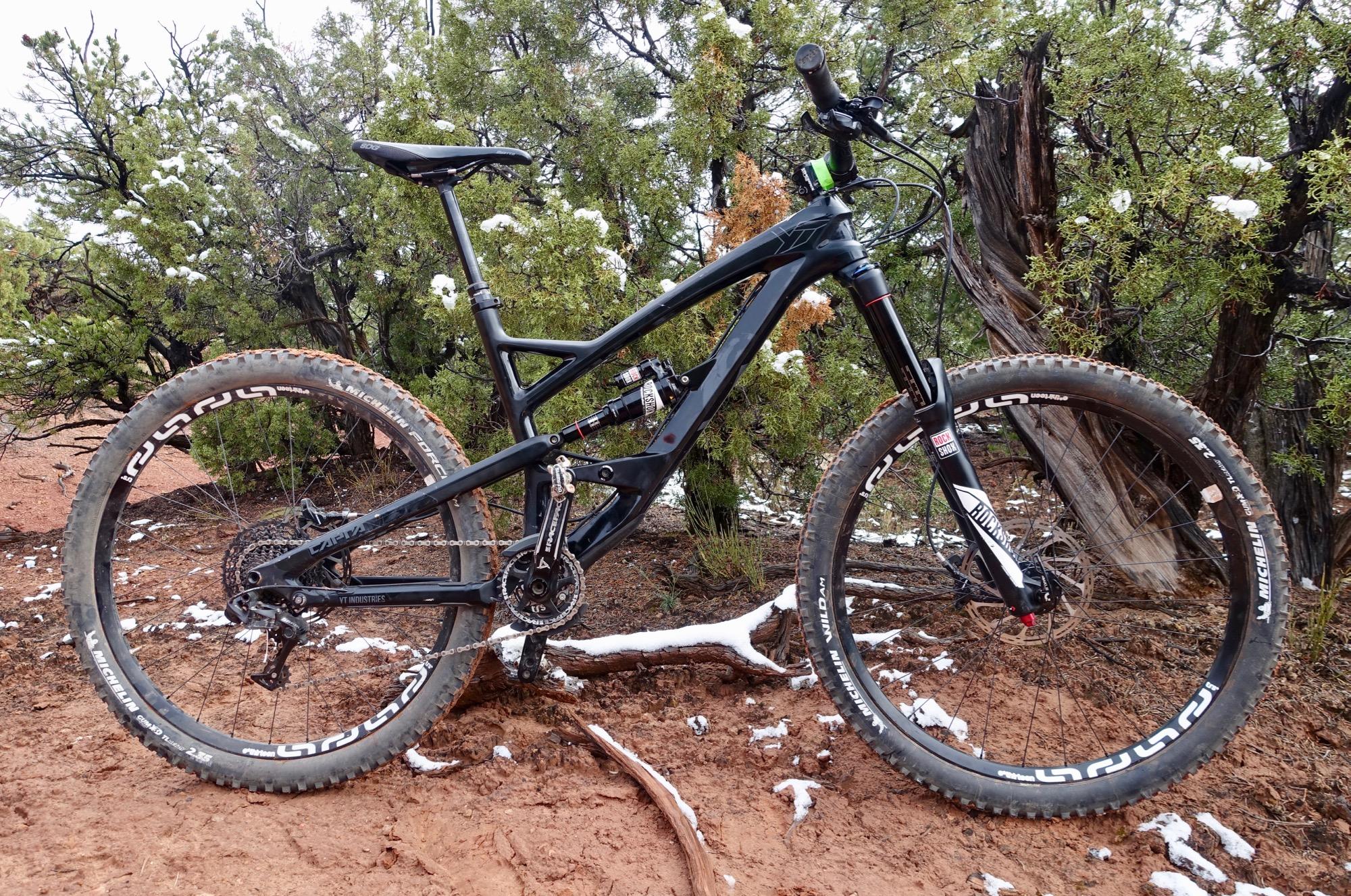 YT Industries Capra Carbon: A black mountain bike standing on a dirt path surrounded by green shrubs and scattered snow. The bike features large tires with visible tread patterns and a high-performance suspension system. The landscape showcases a mix of earthy tones and greenery, indicating a natural outdoor setting.