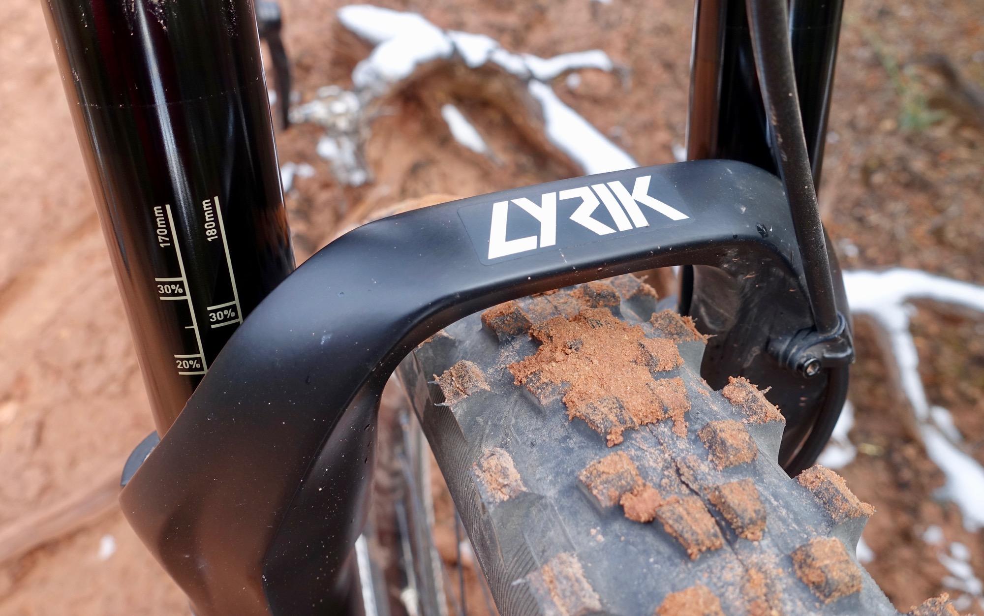 YT Industries Capra Carbon: Close-up of a mountain bike fork labeled "LYRIK" resting on a muddy tire, with visible markings indicating suspension travel measurements (170mm and 180mm) on the fork leg. The background shows earthy terrain with some debris.