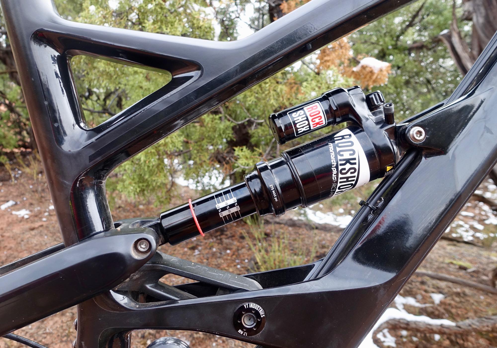 YT Industries Capra Carbon: Close-up view of a bicycle's rear suspension, featuring a RockShox shock absorber mounted within a sleek black frame. The image shows detailed components including a black and red shock with adjustment dials and a YT Industries logo, set against a blurred natural background with greenery and patches of snow.