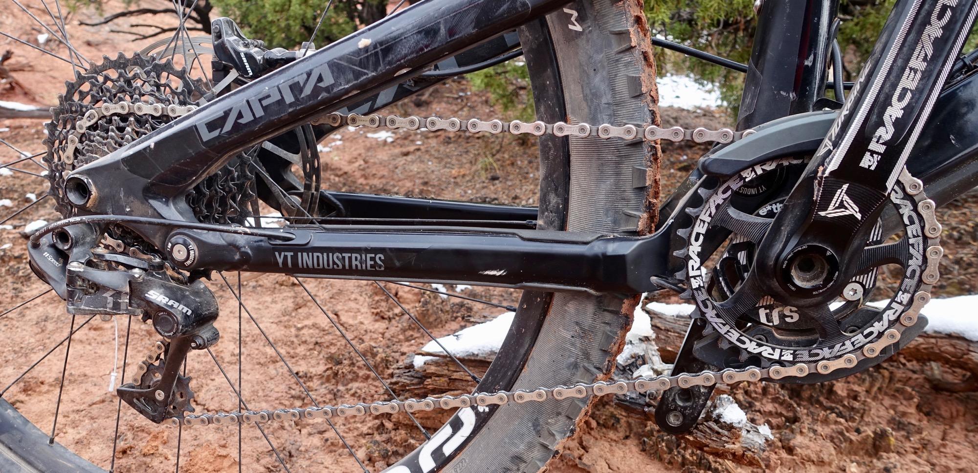 YT Industries Capra Carbon: Close-up view of the rear drivetrain of a mountain bike, featuring a black frame, cassette, and chain. The bike is set against a natural background with dirt and some snow visible, highlighting its rugged terrain suitability. Components are branded with logos from SRAM and YT Industries.