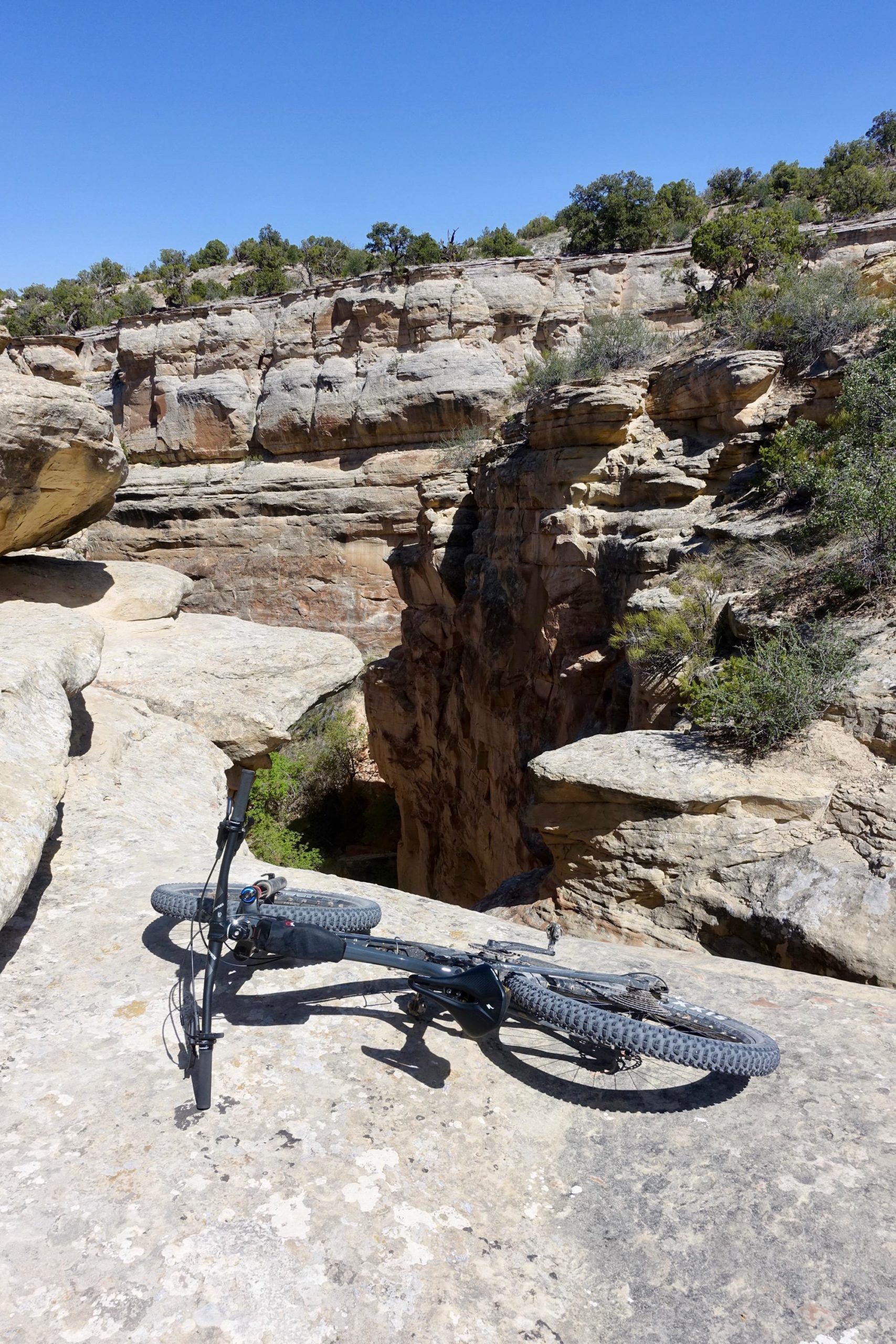 A mountain bike resting on a rocky ledge overlooking a deep canyon under a clear blue sky, surrounded by rugged terrain and sparse vegetation. The Ribbon mountain bike trail.