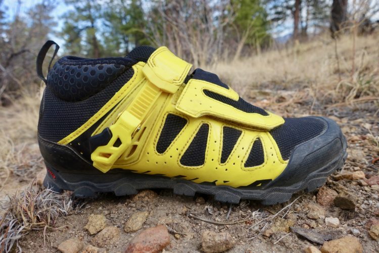 A close-up of a black and yellow athletic shoe designed for outdoor activities, showcasing a textured upper with a prominent closure system and a rugged sole, resting on a natural, rocky surface with sparse vegetation in the background.