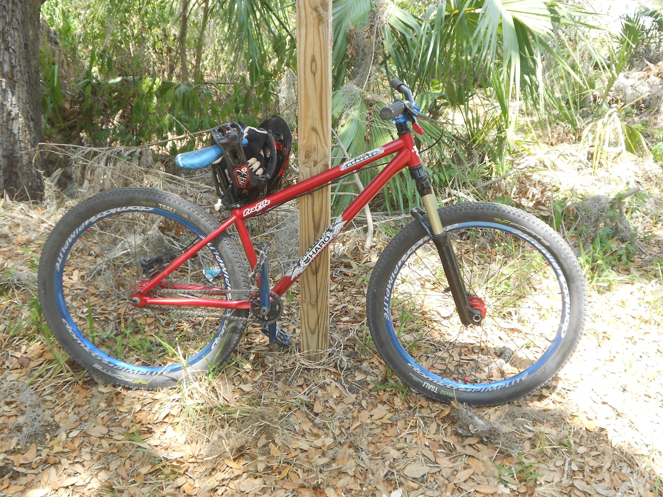 A red mountain bike leaning against a wooden post surrounded by greenery, with a black bag attached to the saddle. The scene includes scattered leaves on the ground and various plants in the background. Grapefruit Trail mountain bike trail.