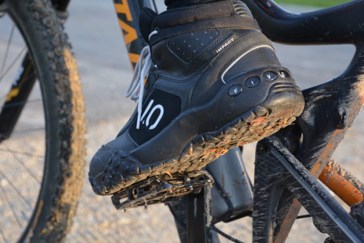 Close-up image of a black sneaker with the word "impact" visible, resting on a bicycle pedal. The shoe features a textured sole for grip and has some dirt smeared on it, suggesting outdoor use. The bicycle frame is partially visible in the background, showcasing its tire and colored details.