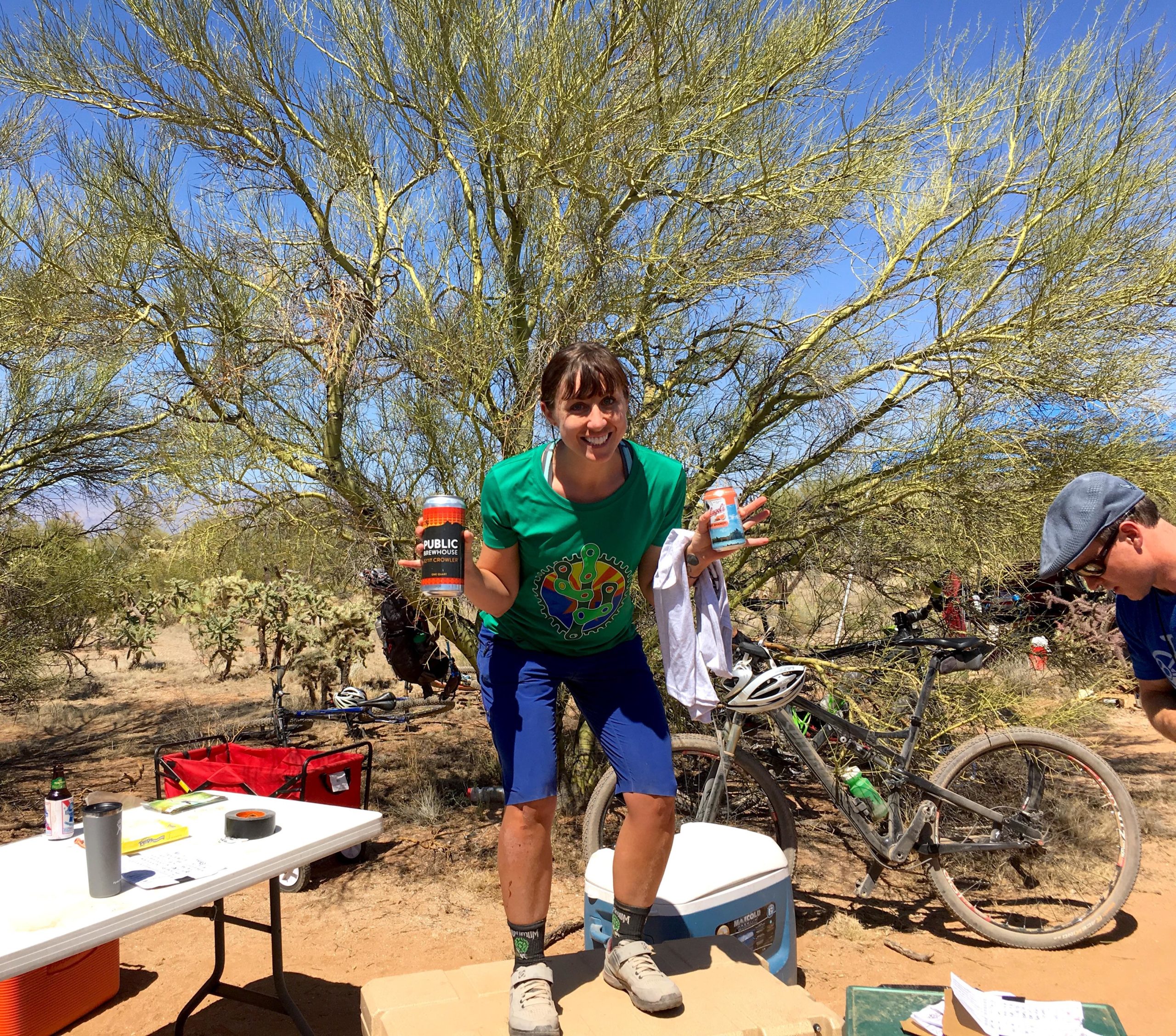 A person wearing a colorful t-shirt and blue shorts smiles while holding two cans, one in each hand, standing on top of a cooler in a desert landscape. In the background, there are leafy trees and a mountain bike leaning against a bush. A table with various items is visible nearby, and another person is seen working at the table. The scene is bright and sunny. Fantasy Island mountain bike trail.