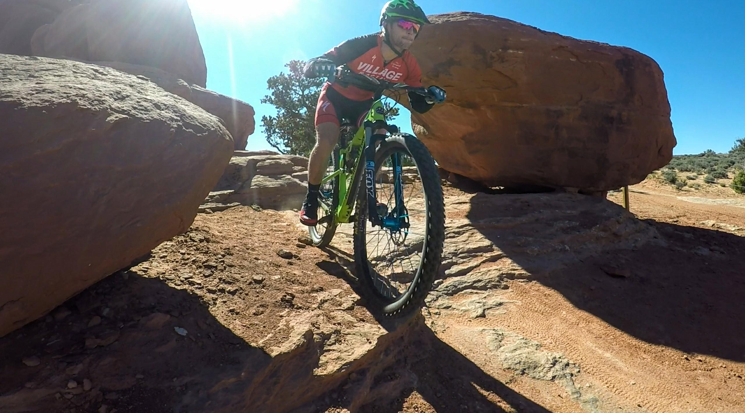 A mountain biker navigates rocky terrain under bright sunlight, showcasing an adventurous outdoor setting. The cyclist is wearing a red jersey and a helmet with vibrant green accents, focused on maneuvering the bike around large boulders. The landscape features a mix of sand, rocks, and sparse vegetation typical of a desert environment. Navajo Rocks mountain bike trail.