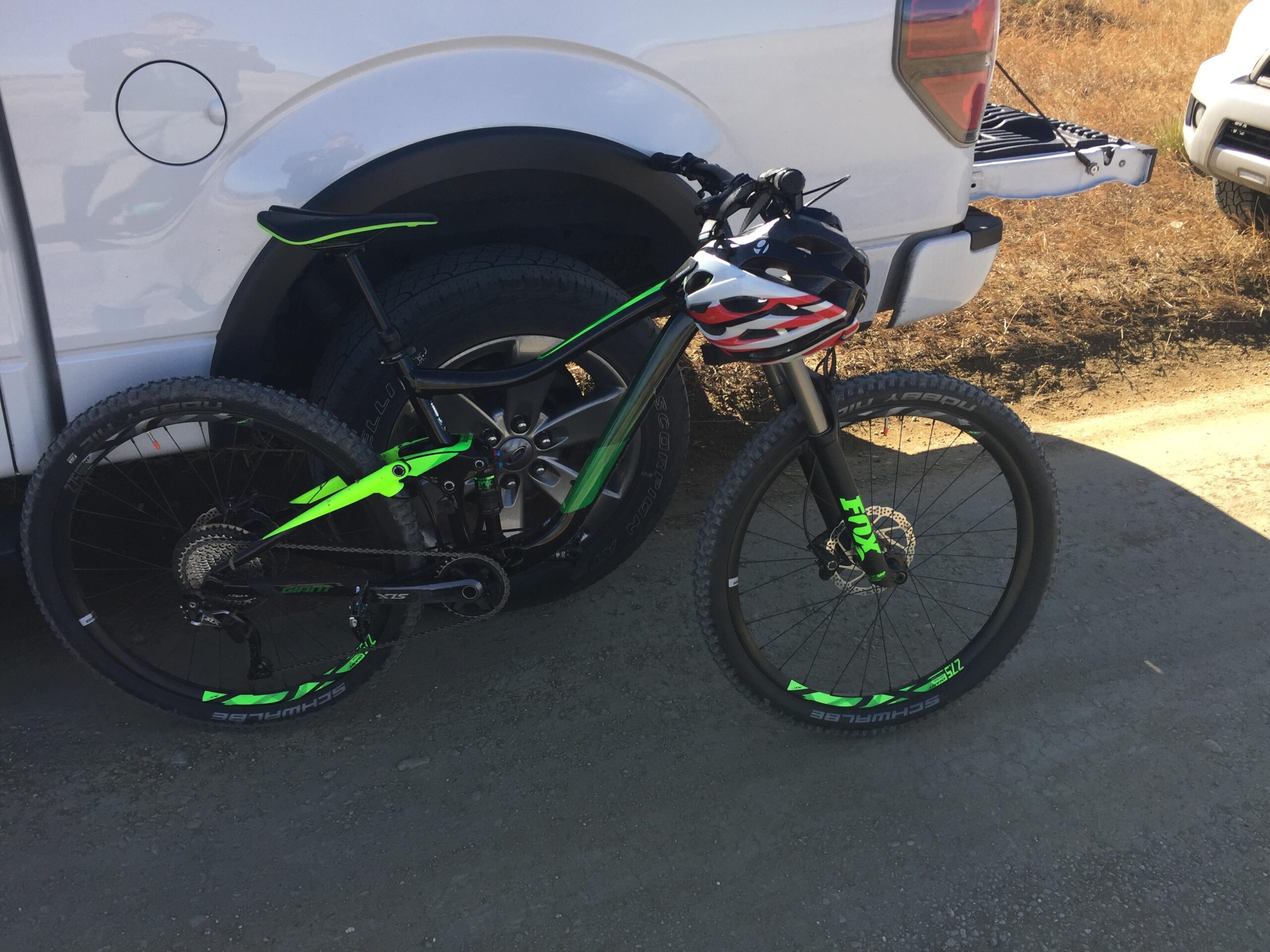 Giant Trance 27.5 2: A mountain bike with a black and green frame is leaning against a white truck. The bike is equipped with thick tires and features a black helmet with a red, white, and blue design placed on its handlebars. The background shows a dirt road and additional vehicles.