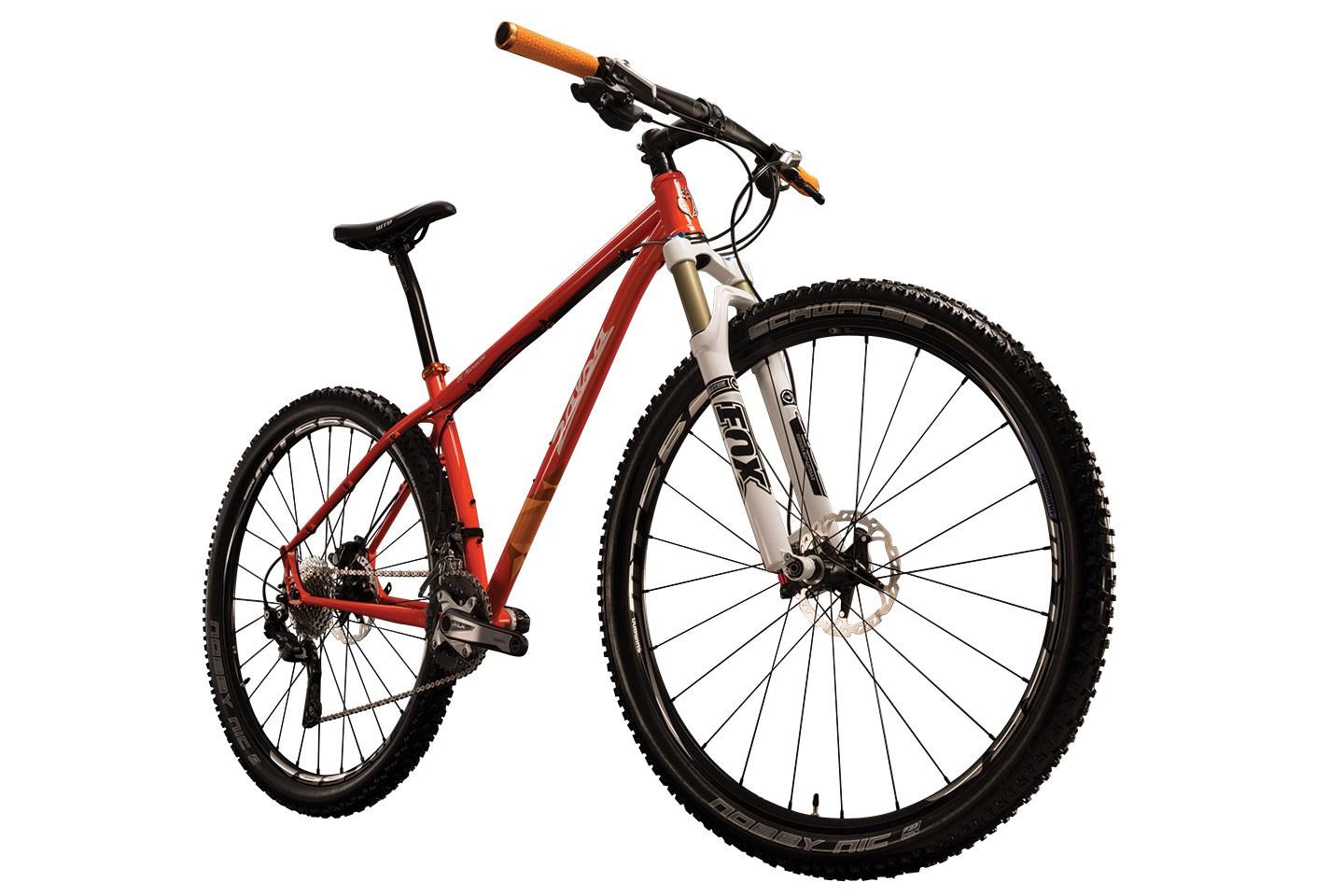 Mountain bike with a red frame, white front fork, and black tires, featuring orange handlebar grips. The bike is positioned at an angle, showcasing its wheels and drivetrain components against a plain white background.
