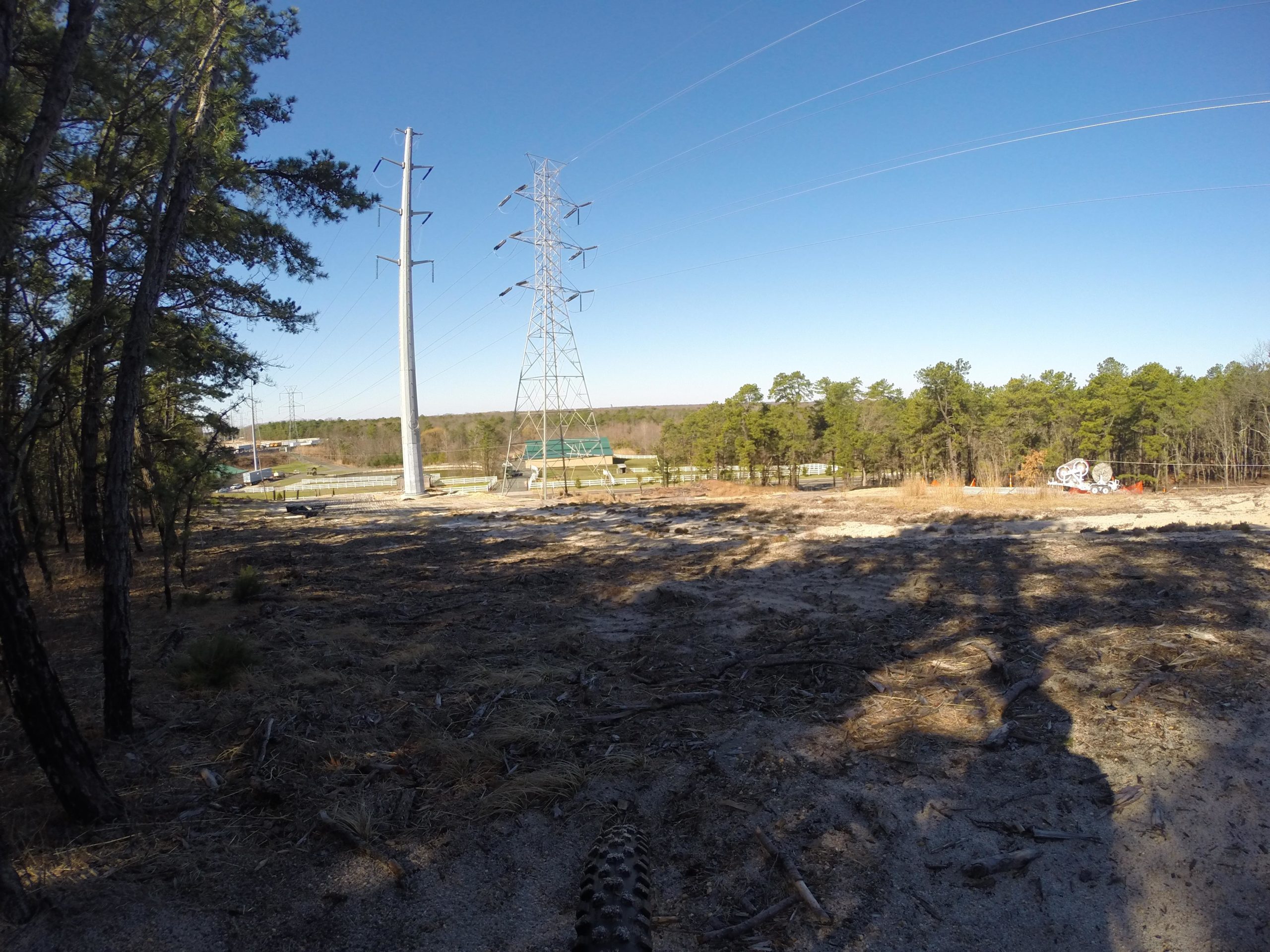 A clear blue sky frames a landscape featuring tall power lines and cleared land. In the foreground, there is a mixture of dirt and sparse vegetation, with tree trunks visible on the left. The background shows a green structure near the base of the power lines, with additional trees and a hint of developed land beyond. Allaire State Park mountain bike trail.