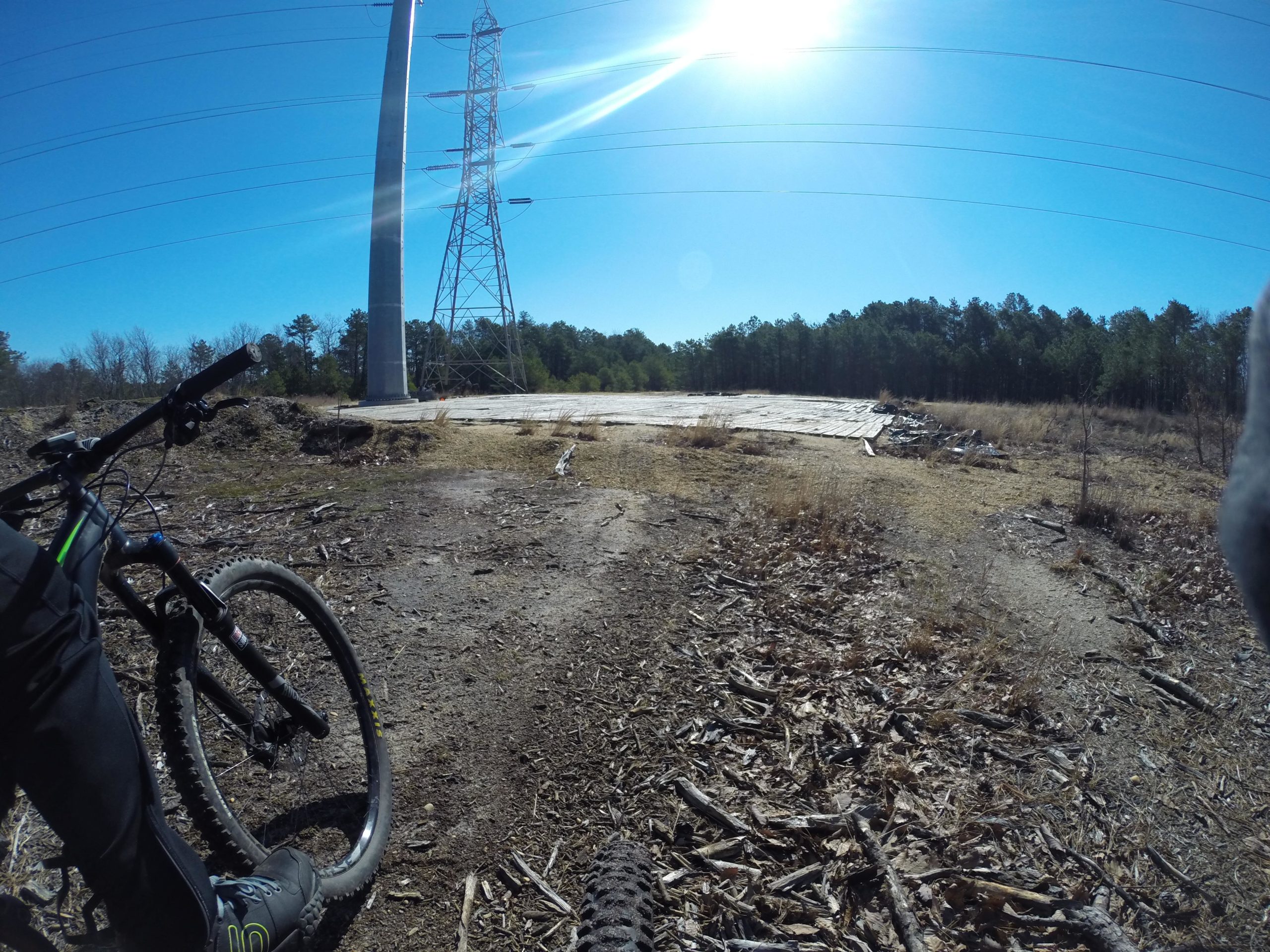 A mountain bike resting on a dirt trail beside a power line tower, with clear blue skies and a sun flare in the upper right corner, surrounded by sparse vegetation and trees in the background. Allaire State Park mountain bike trail.