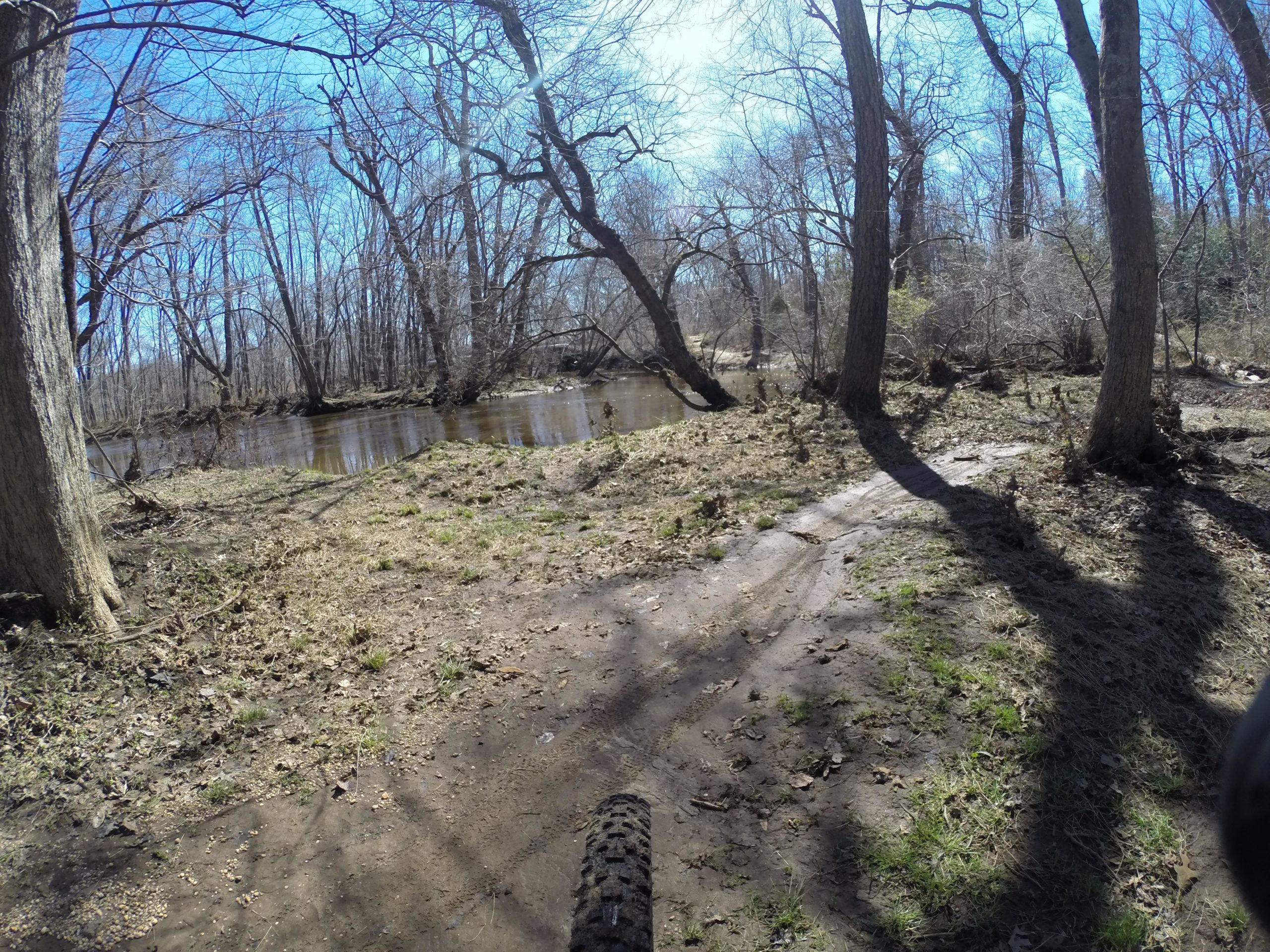 A path winding through a wooded area next to a calm river, with bare trees and a clear blue sky overhead. The ground is covered in a mix of mud and grass, indicating recent activity. Allaire State Park mountain bike trail.