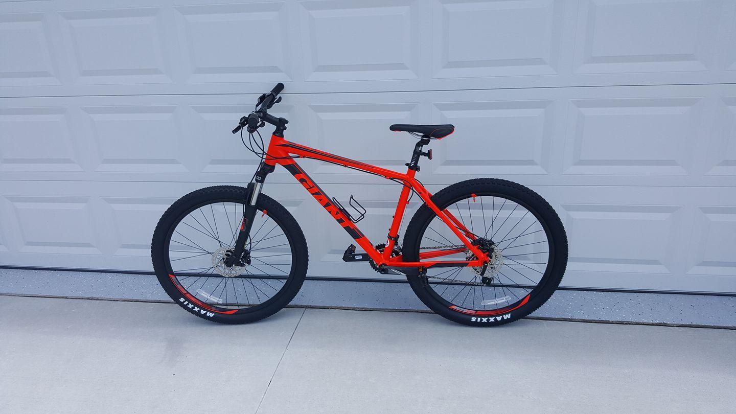 2017 Giant Talon 3: A vibrant orange mountain bike with thick black tires, positioned against a gray garage door. The bike features a sturdy frame, disc brakes, and a water bottle holder, showcasing its readiness for outdoor adventures.