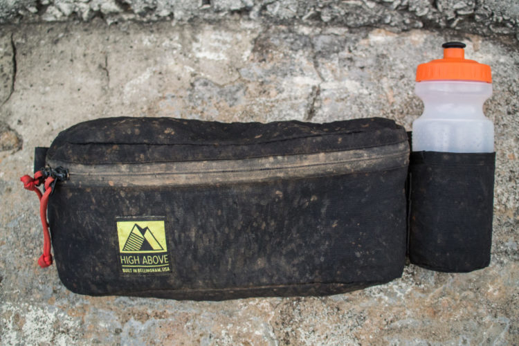 A black, slightly dirty backpacking pouch featuring a yellow "High Above" logo, accompanied by a clear water bottle with an orange lid. The pouch is laid against a textured, rocky background.