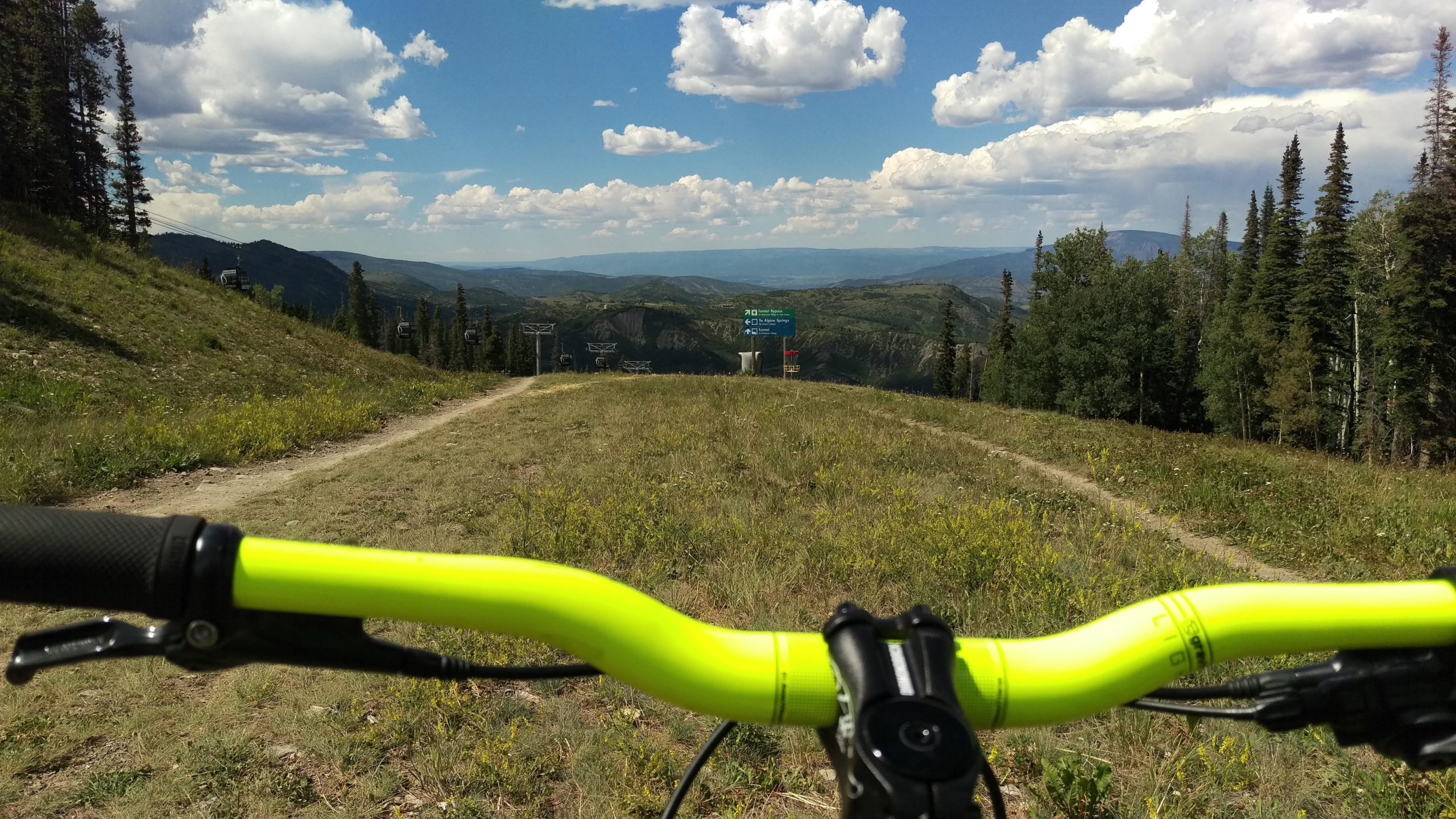 A view from the handlebars of a mountain bike, showcasing a grassy trail leading downhill. In the distance, cable car towers are visible against a backdrop of rolling hills and a partly cloudy sky. The bright yellow handlebars contrast with the natural landscape. Bike Snowmass Bike Park mountain bike trail.