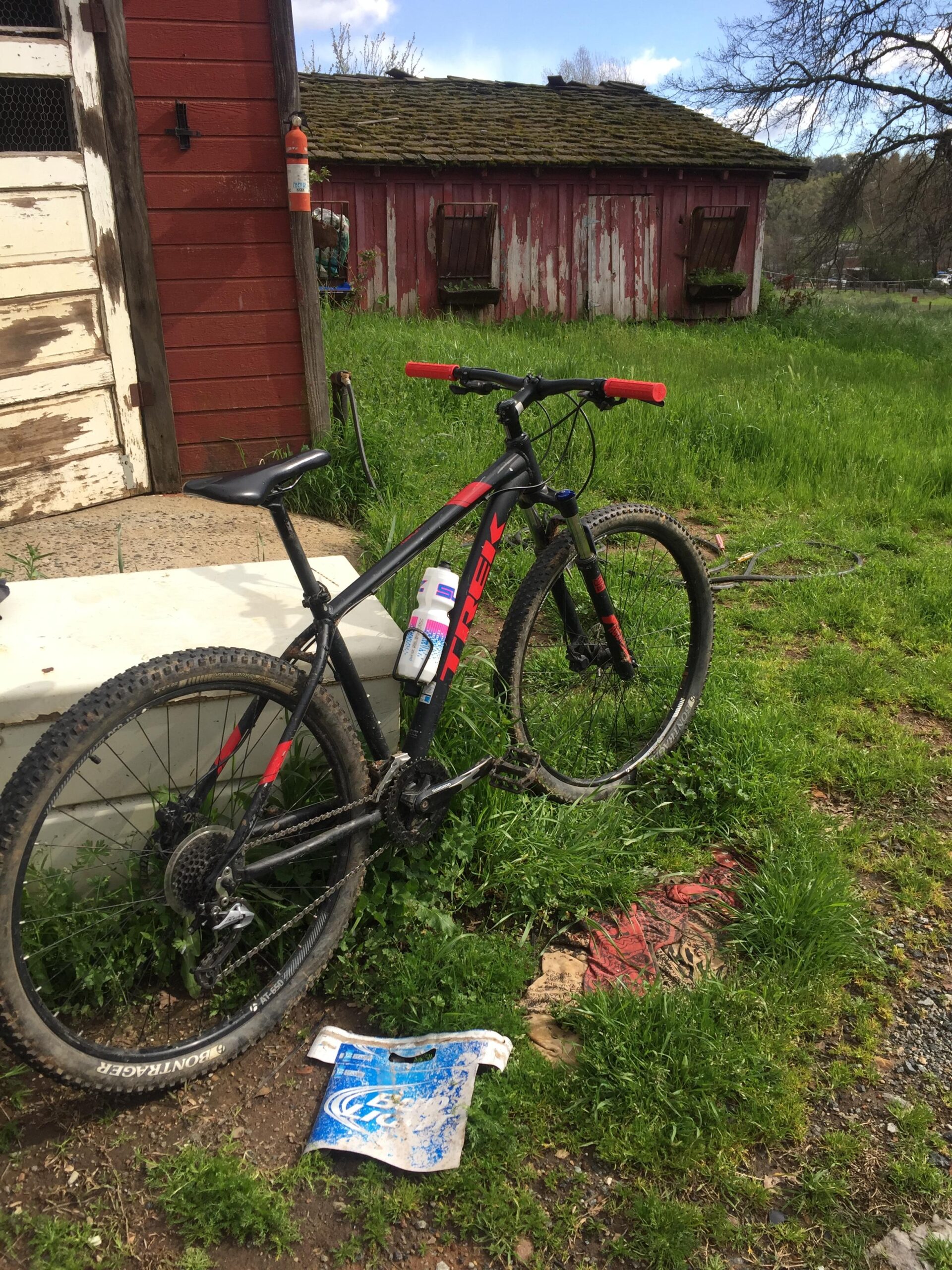 Trek 2017 Trek Marlin 7: A black and red mountain bike rests on a grassy patch next to a rustic red barn. The bike has a water bottle attached to it and shows signs of dirt from use. In the foreground, a soiled piece of blue fabric lies on the ground, blending in with the greenery surrounding it. The scene is set in a rural area with a cloudy sky above.