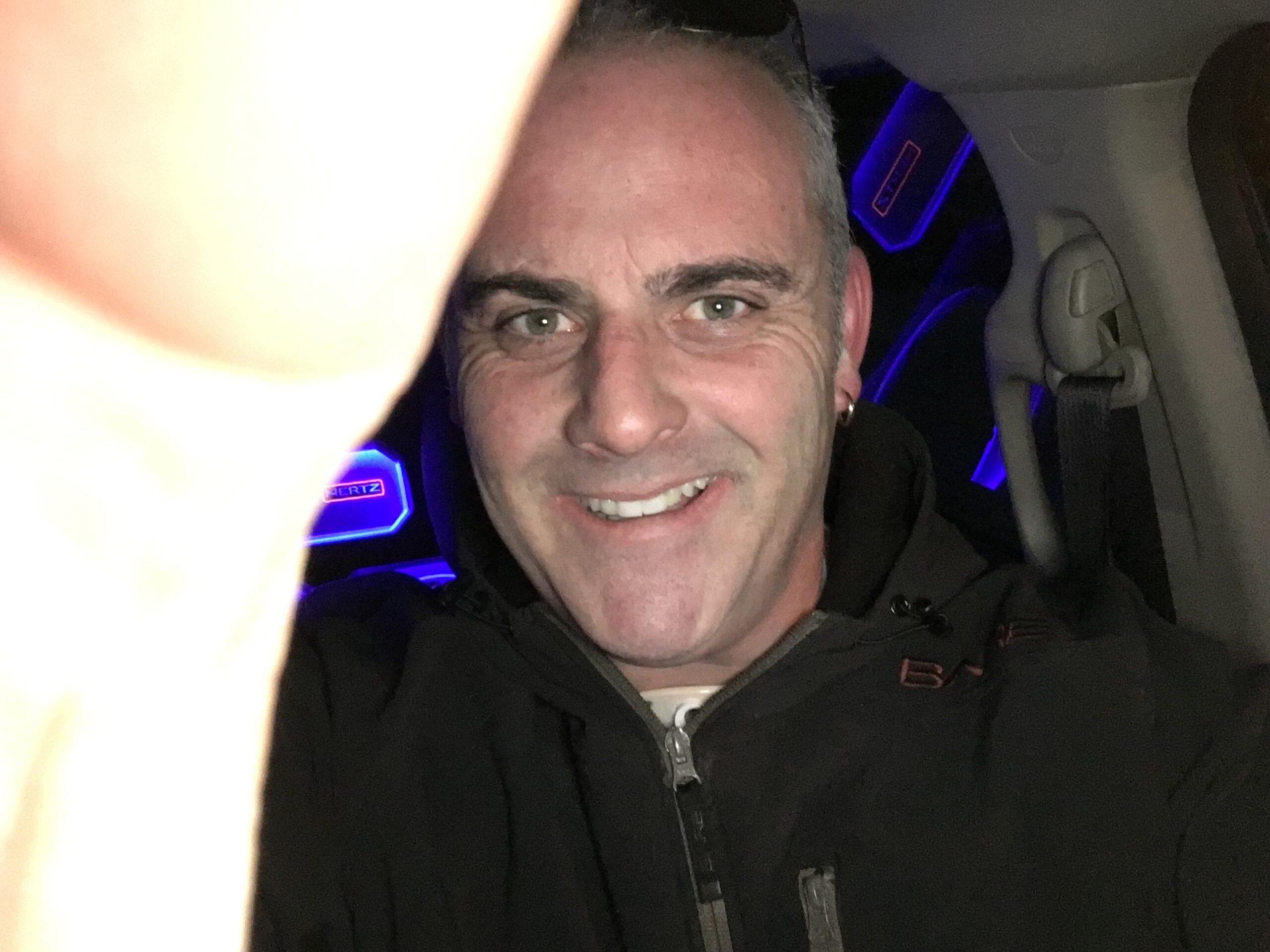 A man smiling at the camera while taking a selfie inside a car, with a partially visible hand in the foreground. The interior features blue ambient lighting. He is wearing a black jacket and has short gray hair.