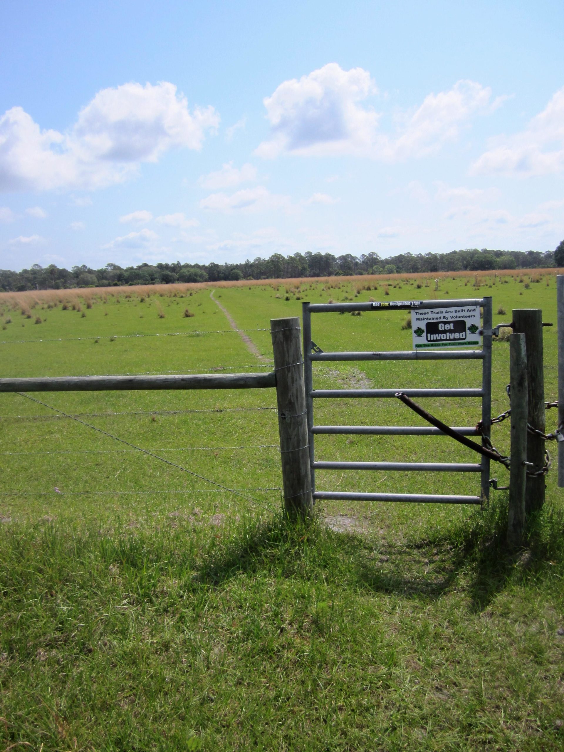 A wooden gate leading into a green, grassy field under a bright blue sky with fluffy white clouds. A sign on the gate encourages people to get involved in volunteer activities related to the trails in the area. In the background, a winding path can be seen moving through the field, lined with sparse vegetation. Grassy Island Trail mountain bike trail.