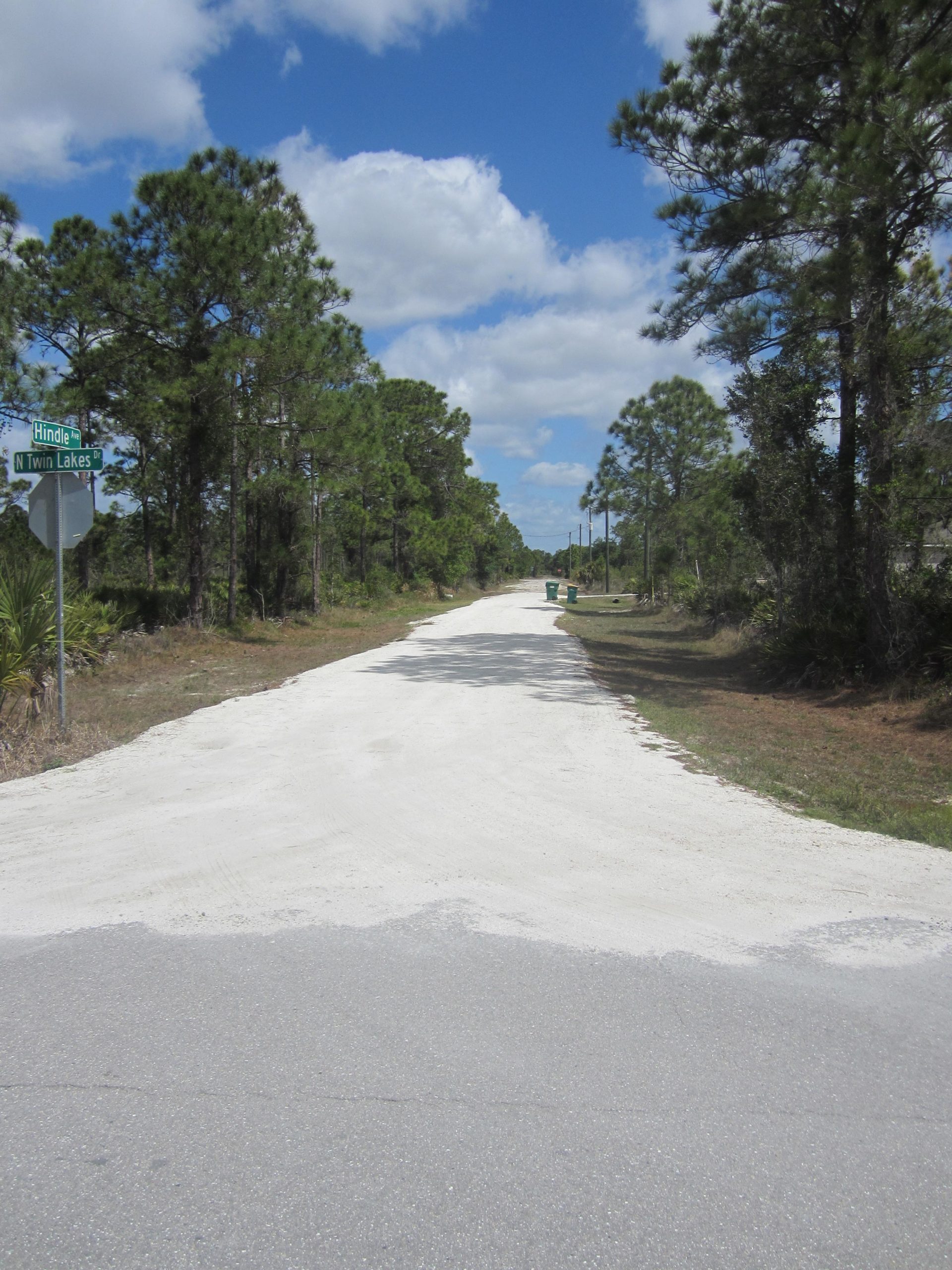 A dirt road lined with trees under a blue sky with fluffy clouds, featuring a street sign indicating "Hindle" and "N Twin Lakes." South County Gravel and Sand mountain bike trail.