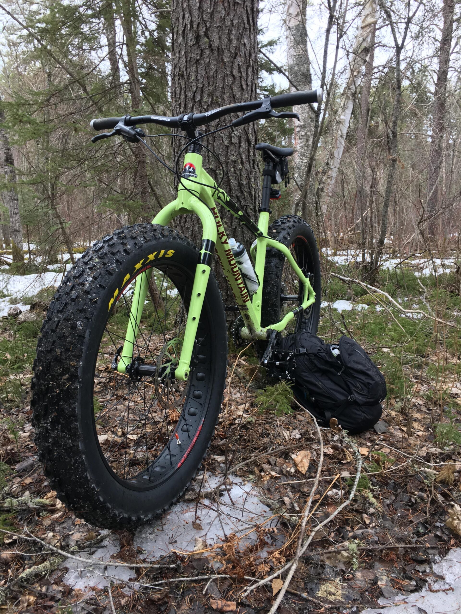 Rocky Mountain Blizzard: A lime green fat bike leaning against a tree in a forested area, with patches of snow and fallen leaves on the ground. A black backpack is positioned near the bike's rear wheel, surrounded by various plants and trees.