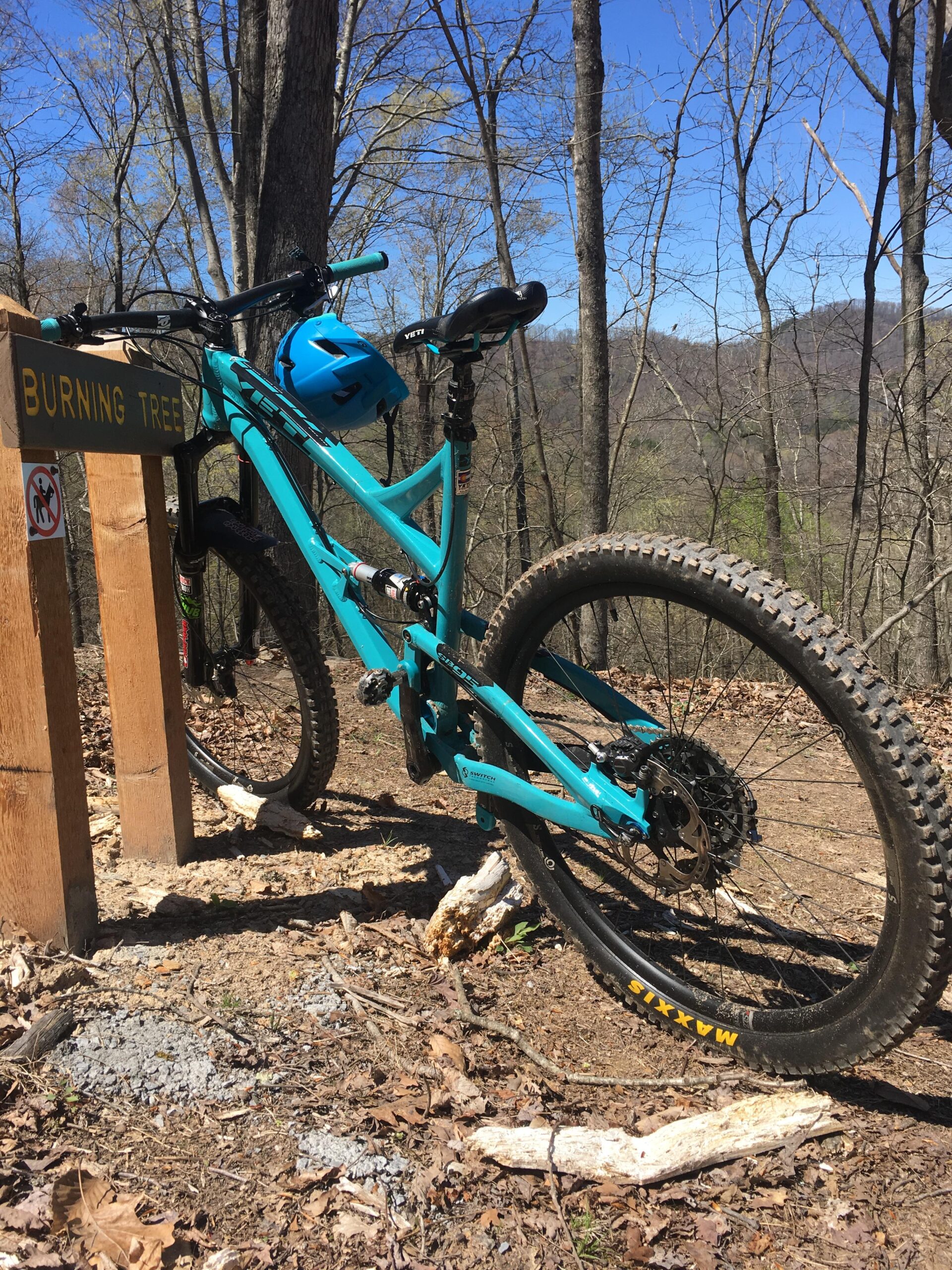 Yeti SB95: A turquoise mountain bike is leaning against a wooden sign that reads "Burning Tree." The bike is equipped with a helmet on the handlebars and is positioned on a dirt trail surrounded by trees. The background features a clear blue sky and distant hills, while the ground is covered with fallen leaves and small branches.