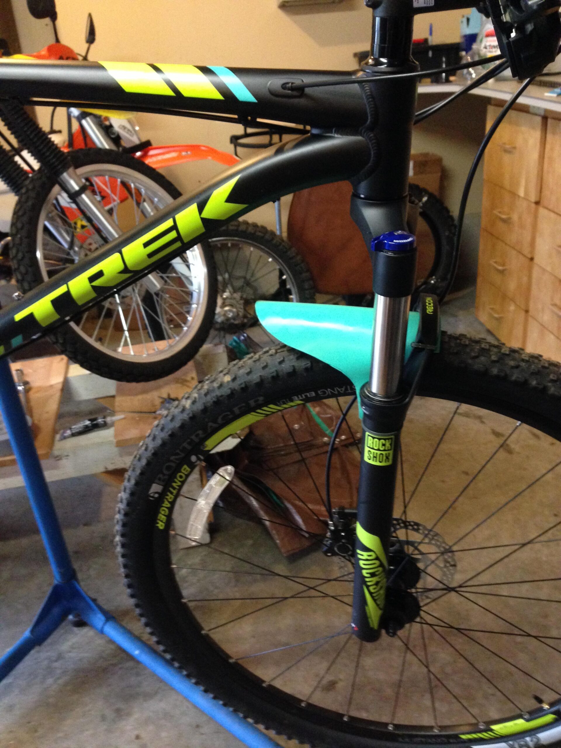 Trek X Caliber 9: A close-up view of a black and green Trek mountain bike, featuring a RockShox suspension fork. The bike is mounted on a blue repair stand in a garage setting, with various tools and another bicycle visible in the background. The front wheel showcases thick, knobby tires suitable for off-road terrain.