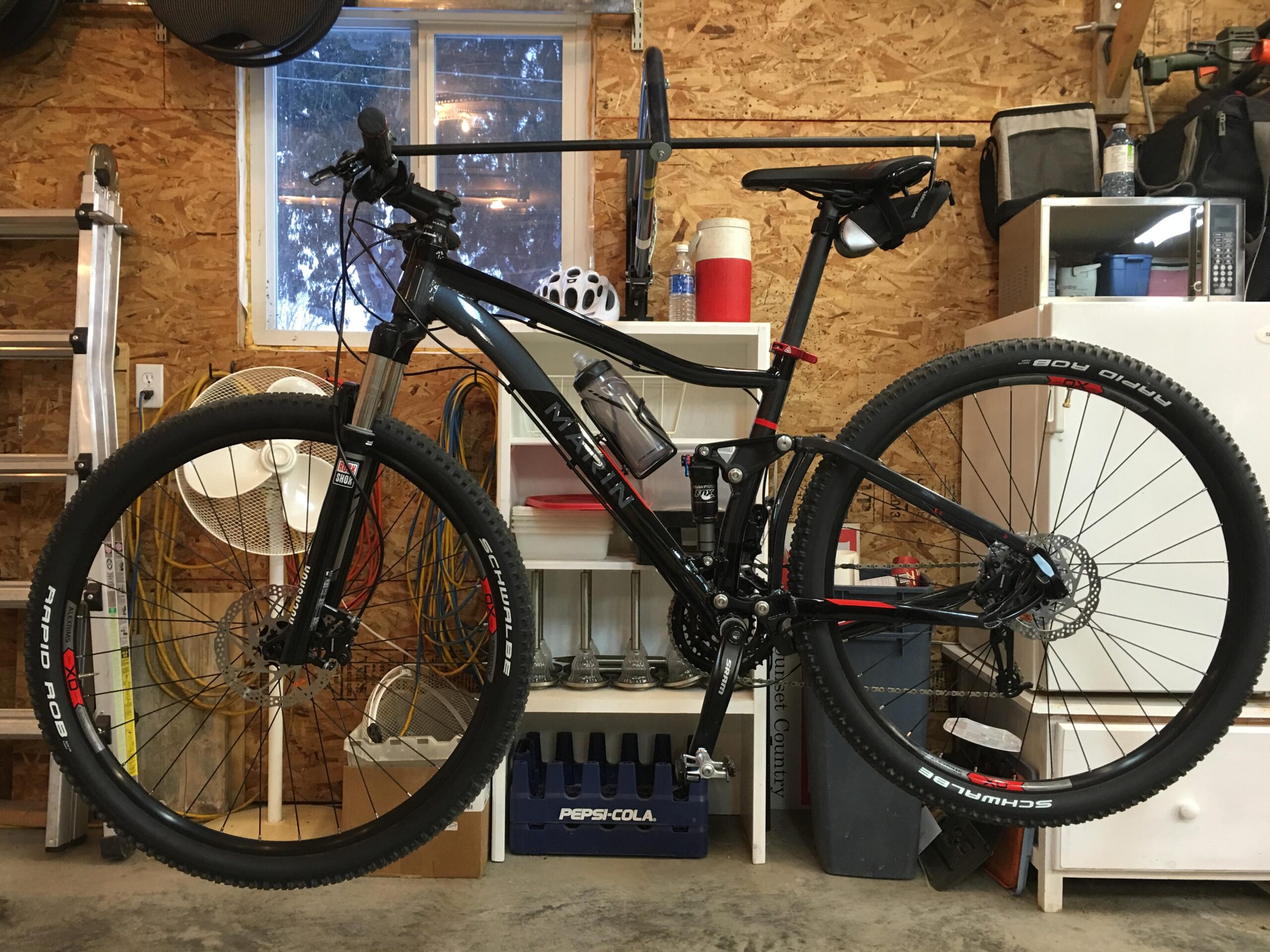 Marin Rift Zone 29er XC6: A mountain bike is hanging in a garage, with a wooden wall in the background. The bike is black with red accents and features thick tires labeled "Schwalbe Rapid Rob." Behind the bike, there are various items including a fan, water bottles, and storage containers. The garage has a warm, inviting atmosphere with natural light coming through a window.