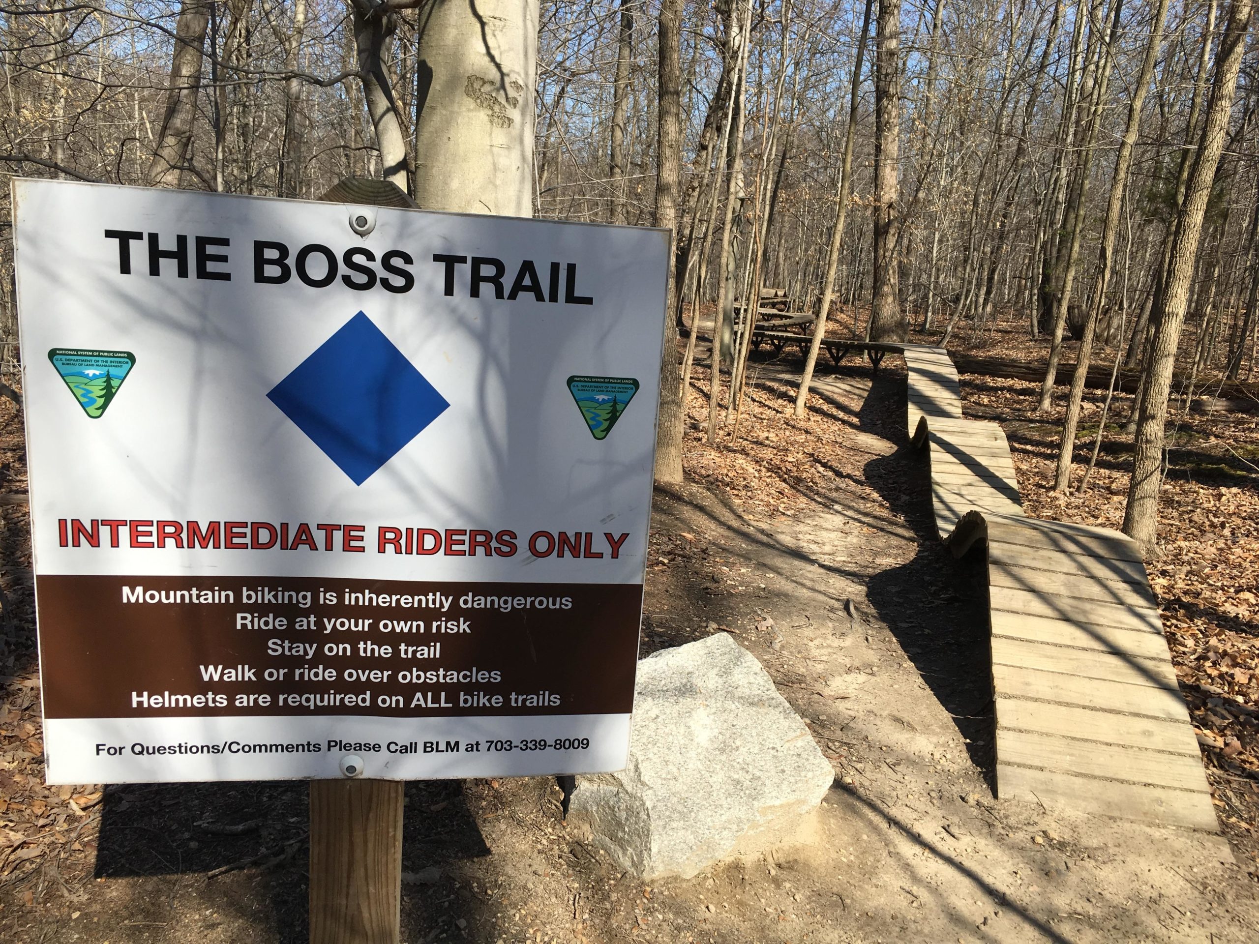 A wooden sign reading "The Boss Trail" hangs on a tree in a forested area. Below the title, the sign indicates that the trail is for "Intermediate Riders Only" and includes safety warnings about mountain biking. Visible trail features include a winding wooden path designed for bikers, surrounded by bare trees and fallen leaves. A stone is partially visible on the trail. Meadowood mountain bike trail.