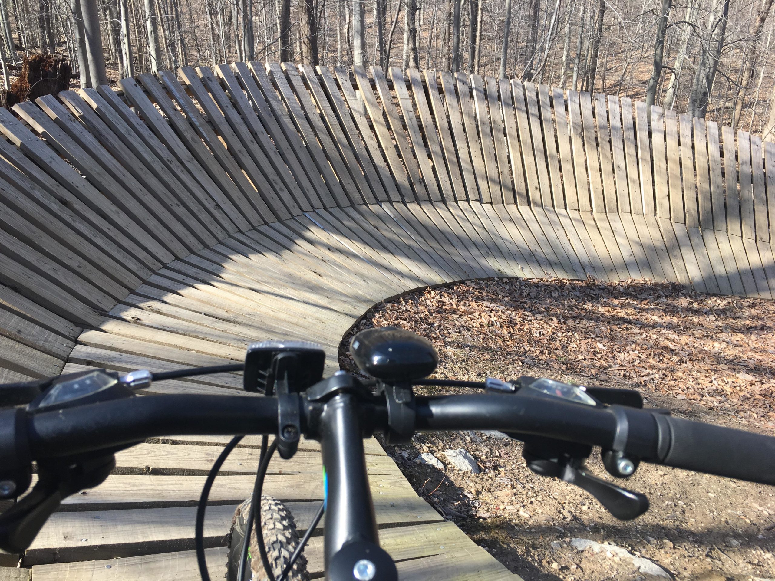 View from the handlebars of a mountain bike approaching a wooden pump track, featuring a series of curved wooden boardwalks surrounded by bare trees and leaves on the ground. Meadowood mountain bike trail.