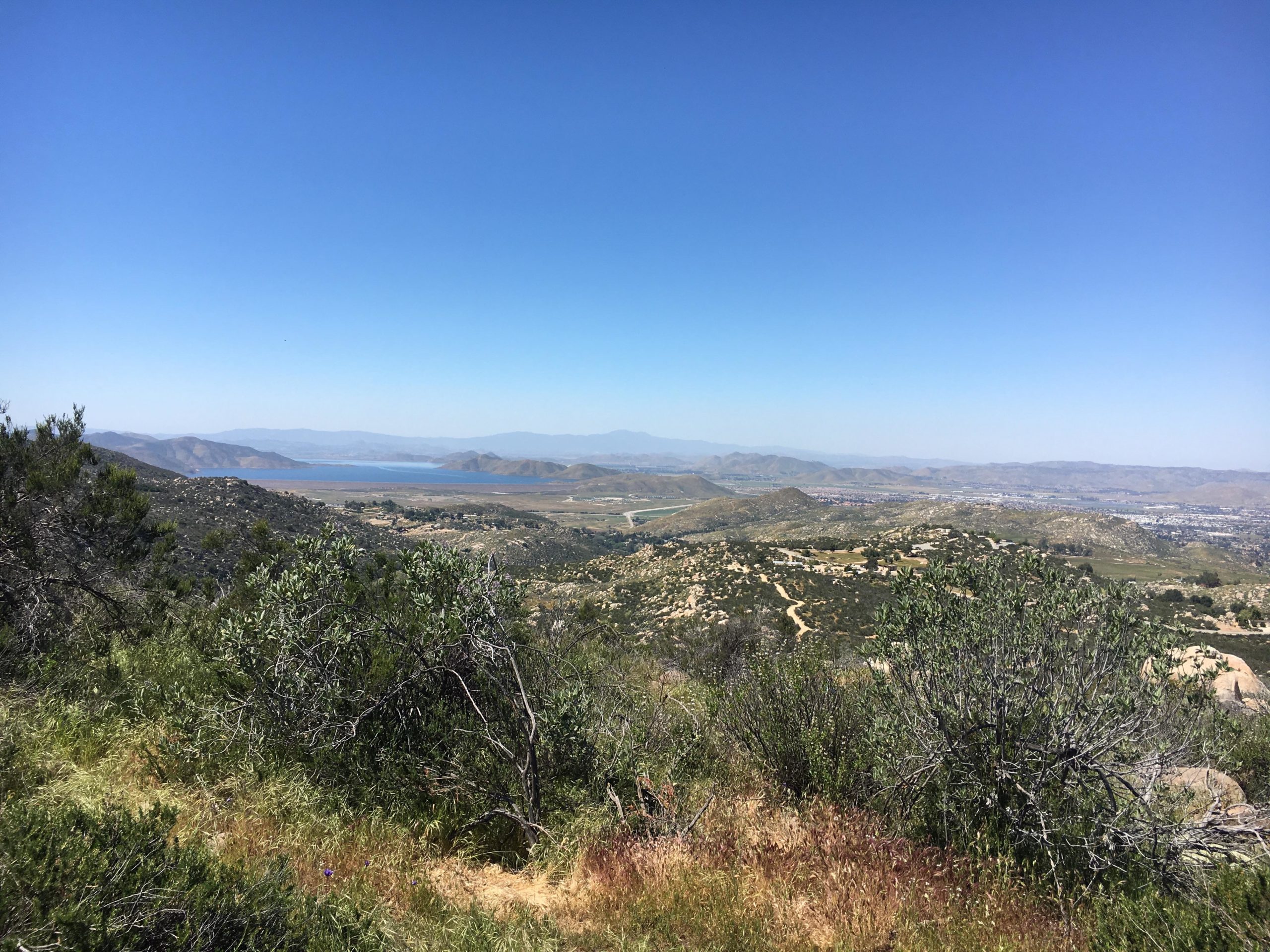 A panoramic view of a hilly landscape under a clear blue sky, featuring patches of greenery and shrubs in the foreground. In the distance, a body of water is visible, surrounded by rolling hills and mountains. Trails and patches of open land indicate a rural setting. Simpson Park mountain bike trail.