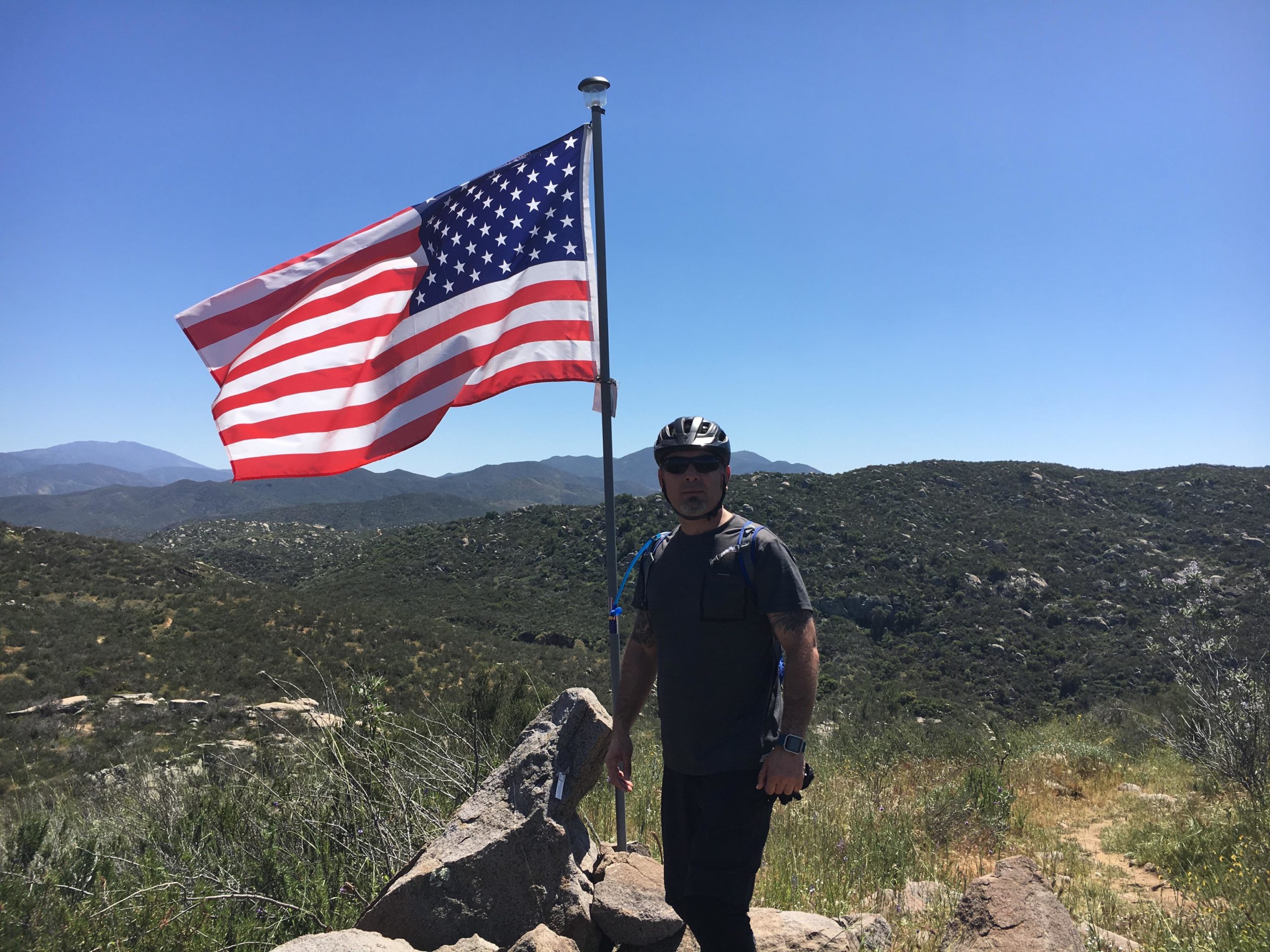 A person wearing a helmet and sunglasses stands beside a tall pole with an American flag waving in the background. The scene is set in a mountainous area with greenery and rocky terrain under a clear blue sky. Simpson Park mountain bike trail.