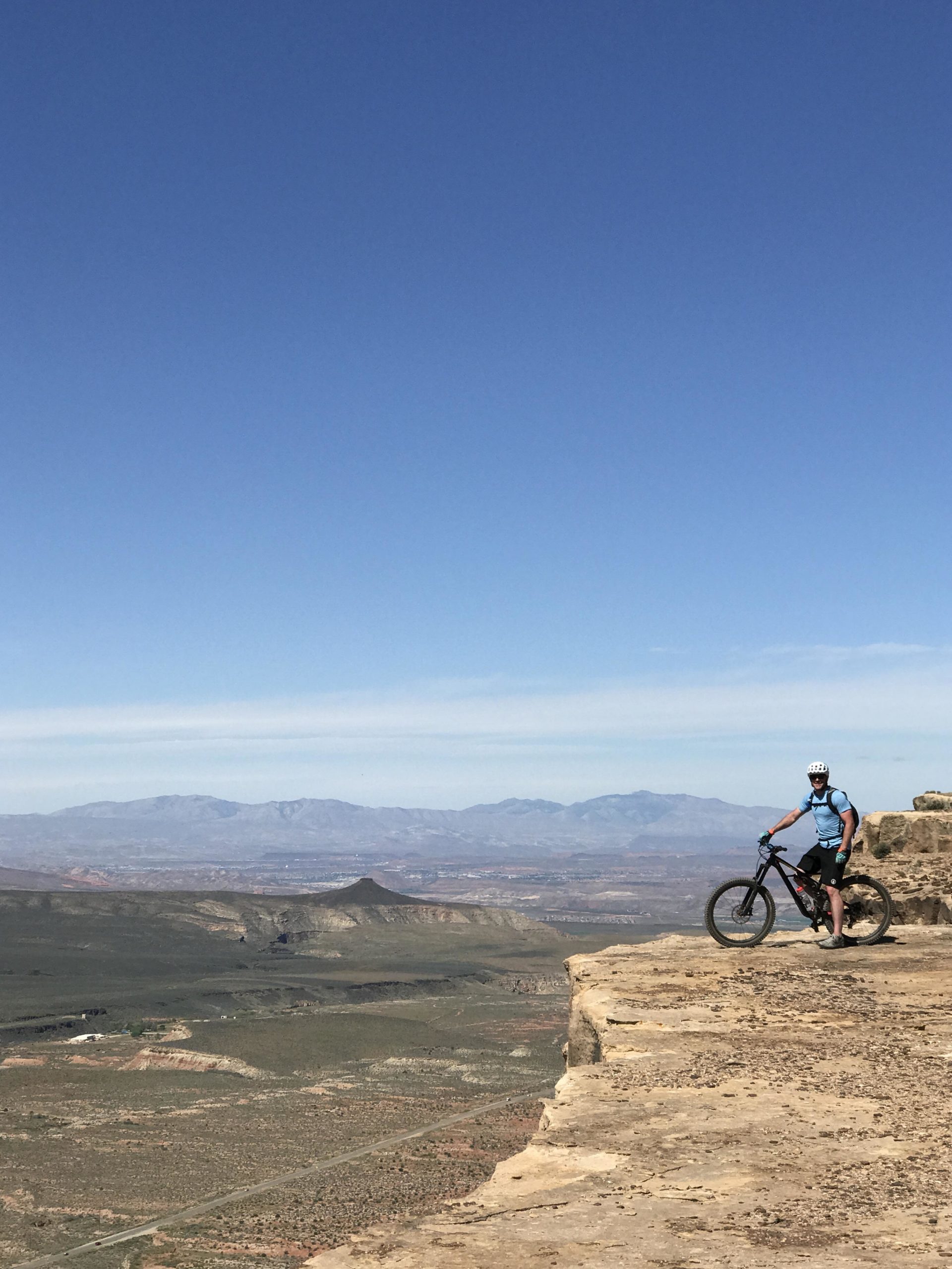 A mountain biker stands on the edge of a rocky cliff, overlooking a vast landscape with rolling hills and distant mountains under a clear blue sky. Gooseberry Mesa mountain bike trail.