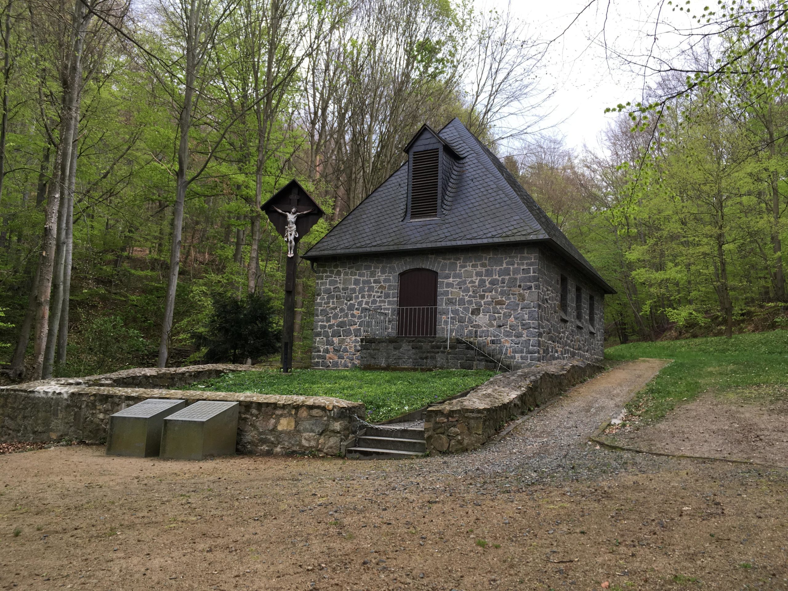A stone building with a dark slate roof situated in a green forest. In front of the building, there is a statue of a crucifix on a wooden post and two metallic benches. The surrounding area features lush green trees and a pathway leading away from the structure. Noerdliche Bergstraße mountain bike trail.
