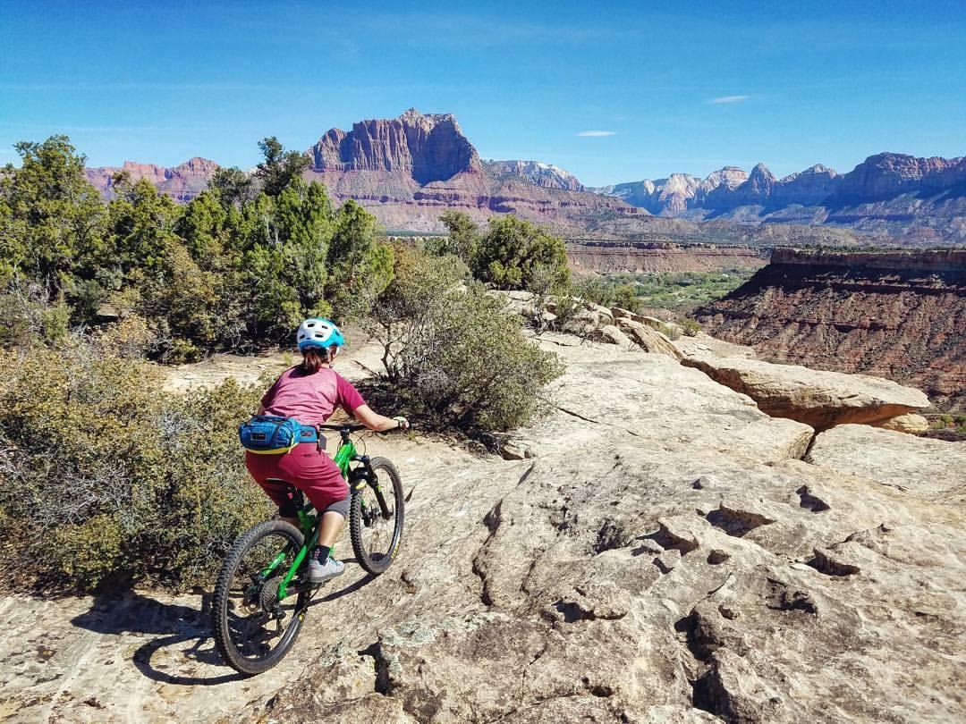 A person riding a mountain bike on rocky terrain, surrounded by green shrubs and a vast landscape featuring red rock formations and distant mountains under a clear blue sky. The cyclist wears a blue helmet and a maroon outfit as they navigate the rugged trail. Wire Mesa Loop mountain bike trail.