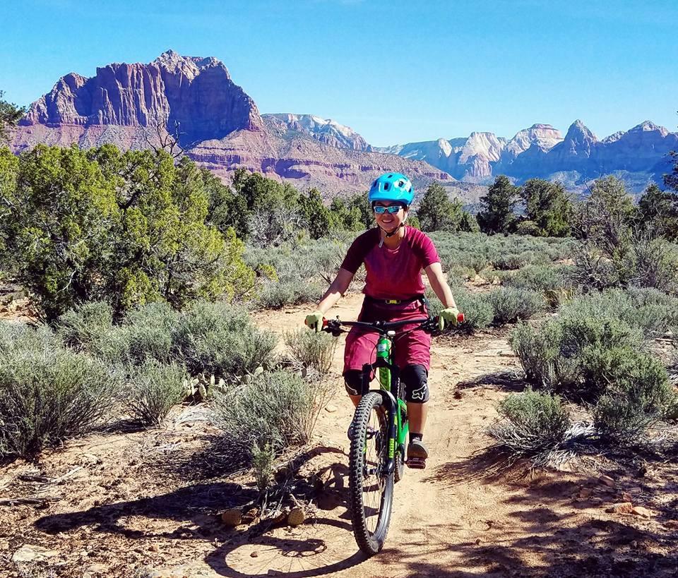 A person riding a mountain bike on a dirt path surrounded by shrubs and small plants, with a backdrop of mountains under a clear blue sky. The cyclist is wearing a blue helmet and sunglasses, along with biking attire in maroon and green. Wire Mesa Loop mountain bike trail.